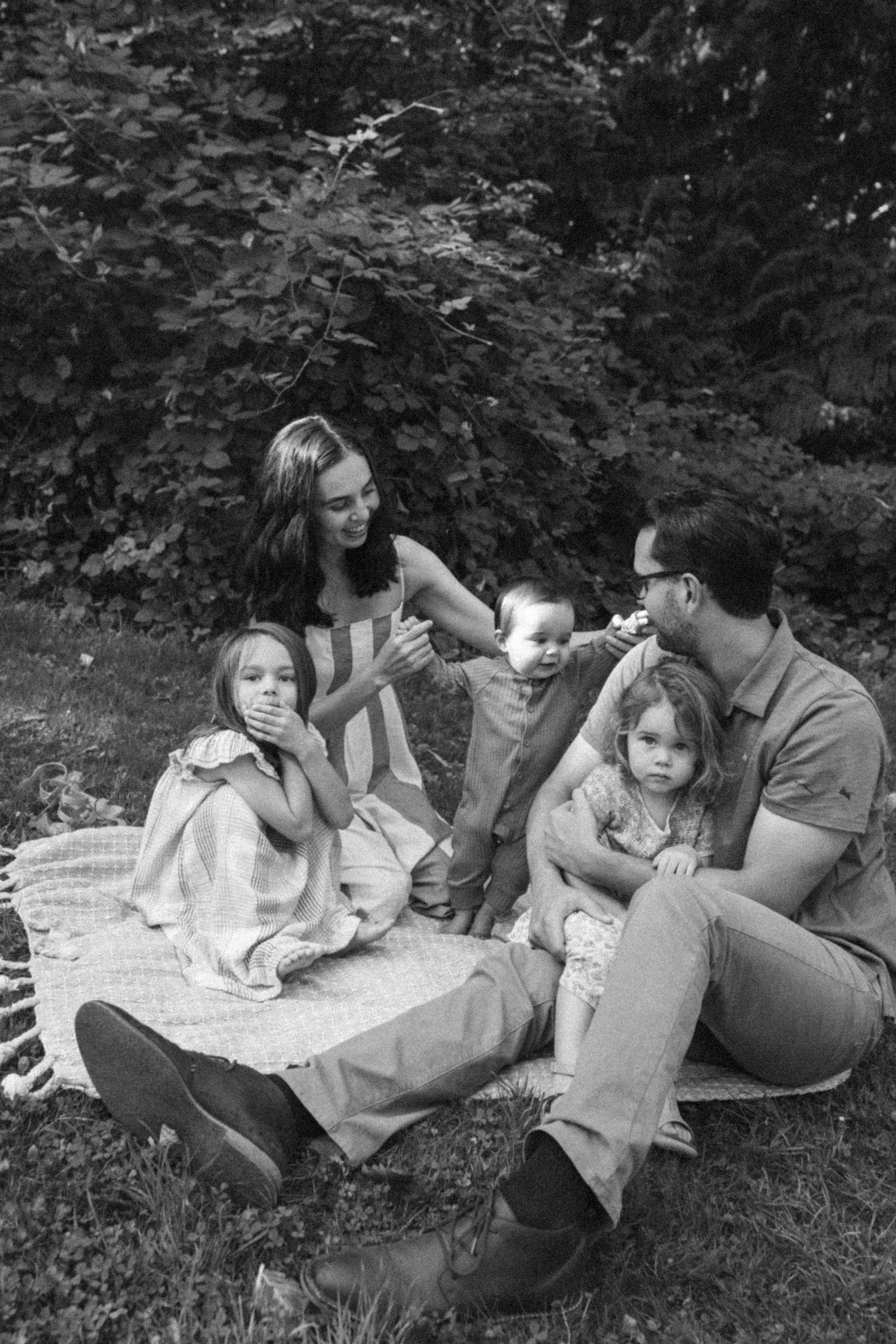 A black and white photo of a family sitting on a blanket outdoors, with trees and foliage in the background. The group includes two adults and three children.