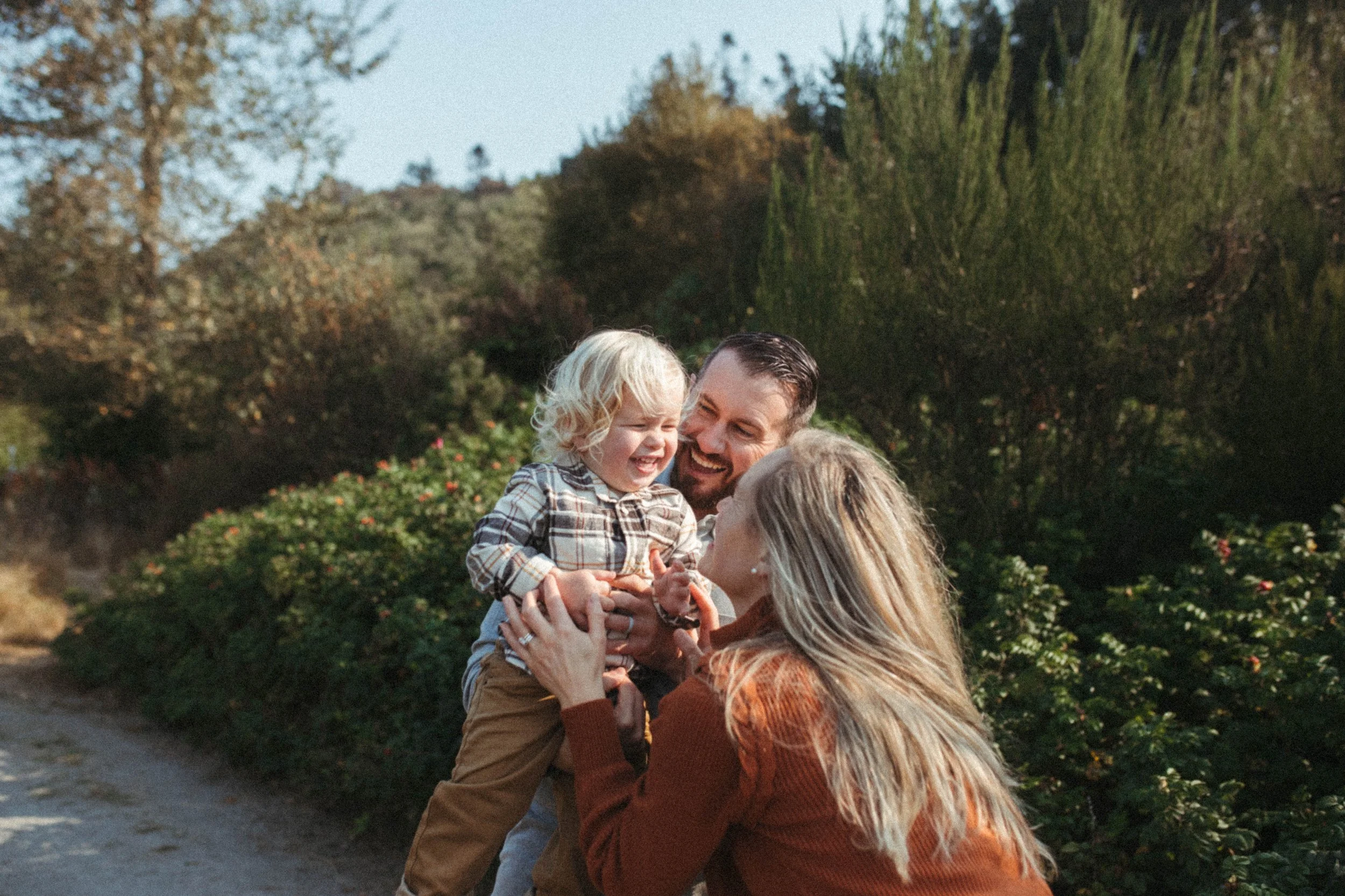 A family of three outdoors, a young boy being held by his mother while his father looks on, all smiling and laughing on a sunny day with trees and bushes in the background.