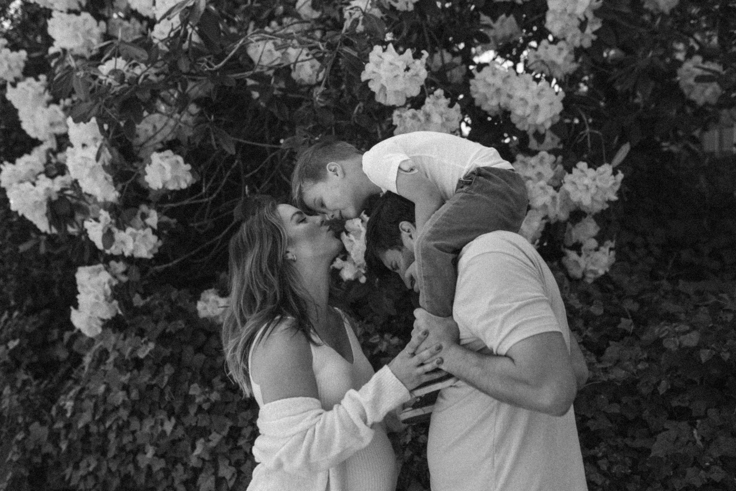A family of three sharing a kiss outdoors. The mother and father hold their young son up as he leans in to kiss her on the nose. They are surrounded by blooming flowers and greenery.