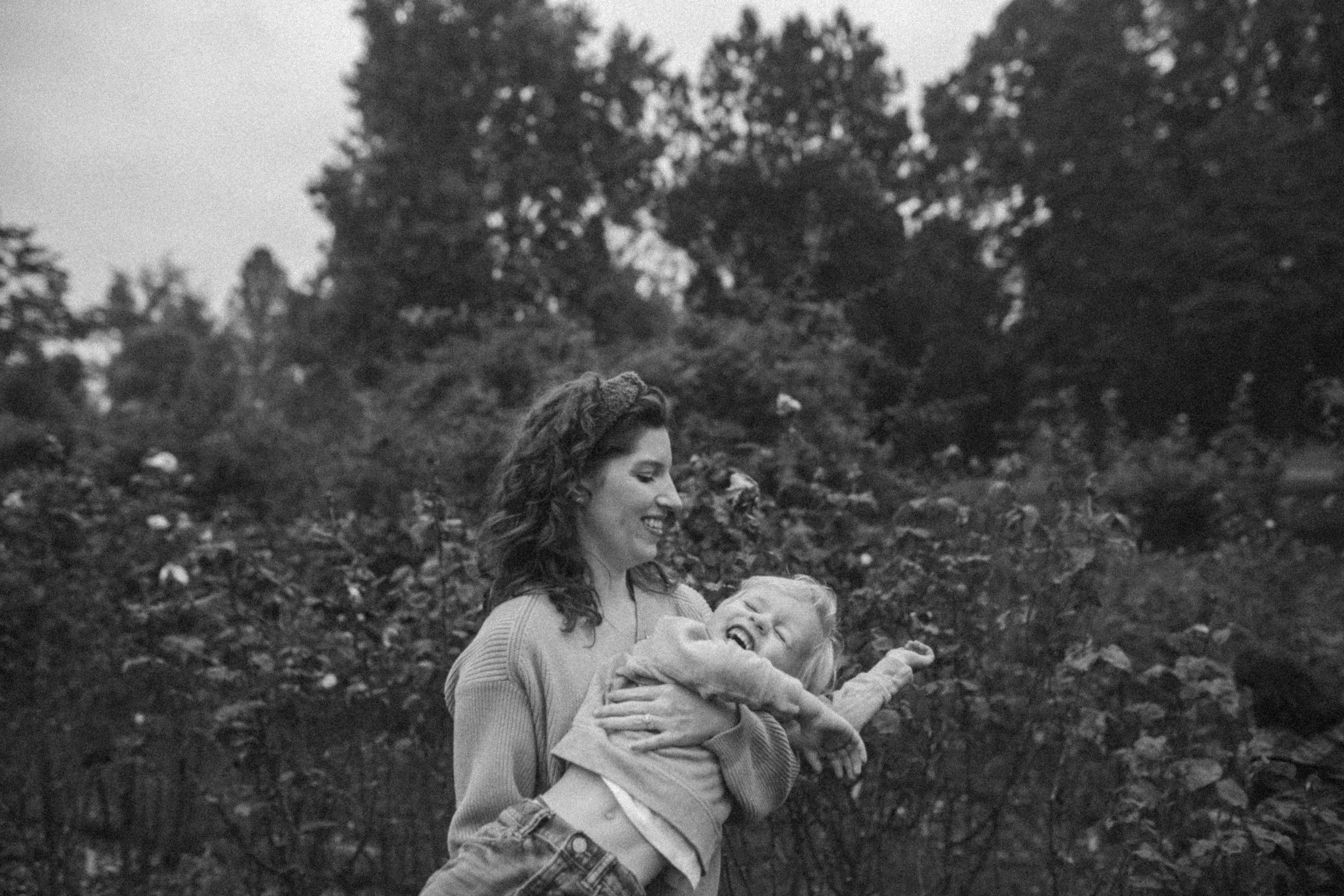 A woman holding a young girl that is laughing outdoors with trees and foliage in the background.