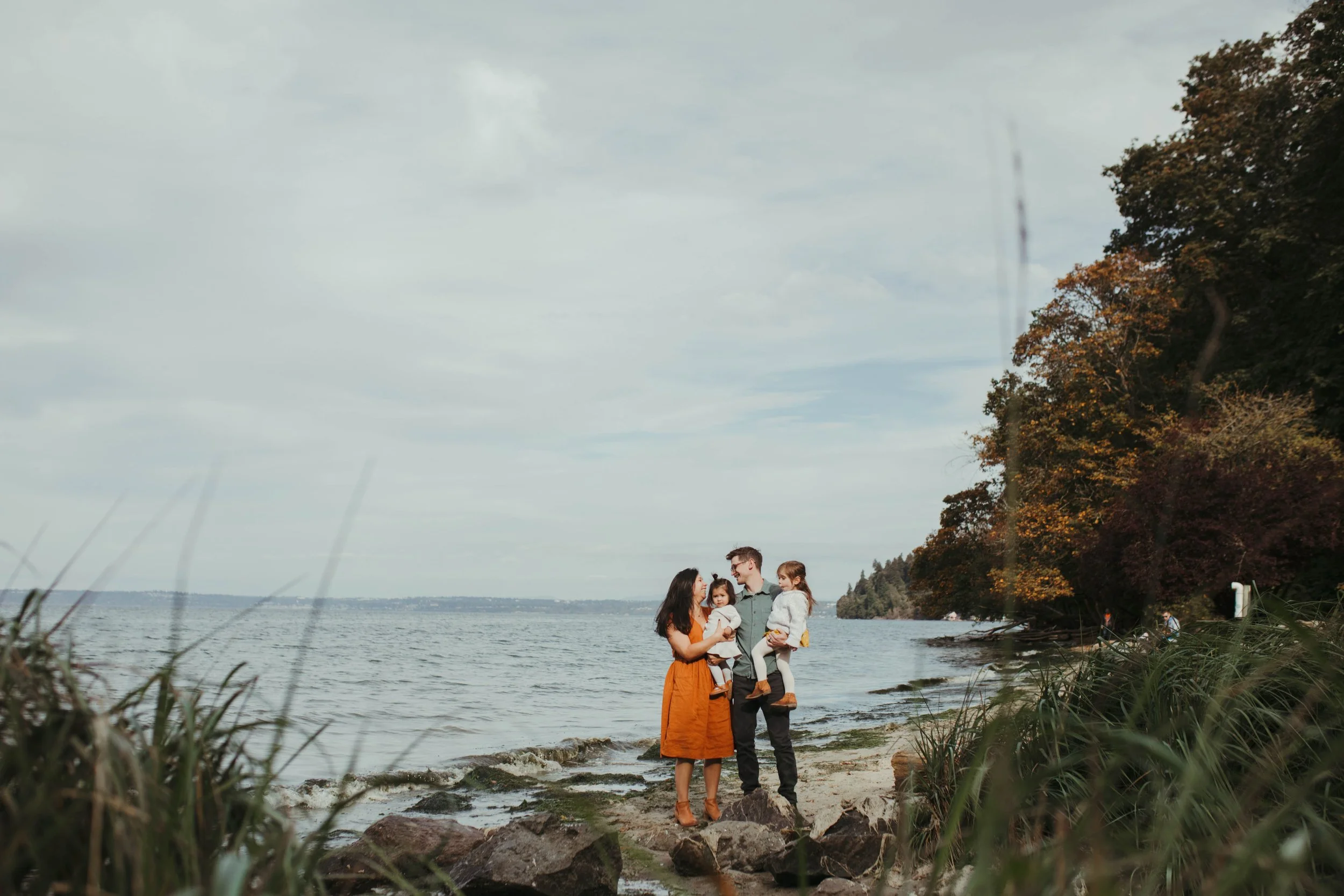 A family of four standing on rocks by a lake, with trees and a cloudy sky in the background, enjoying a day outdoors.