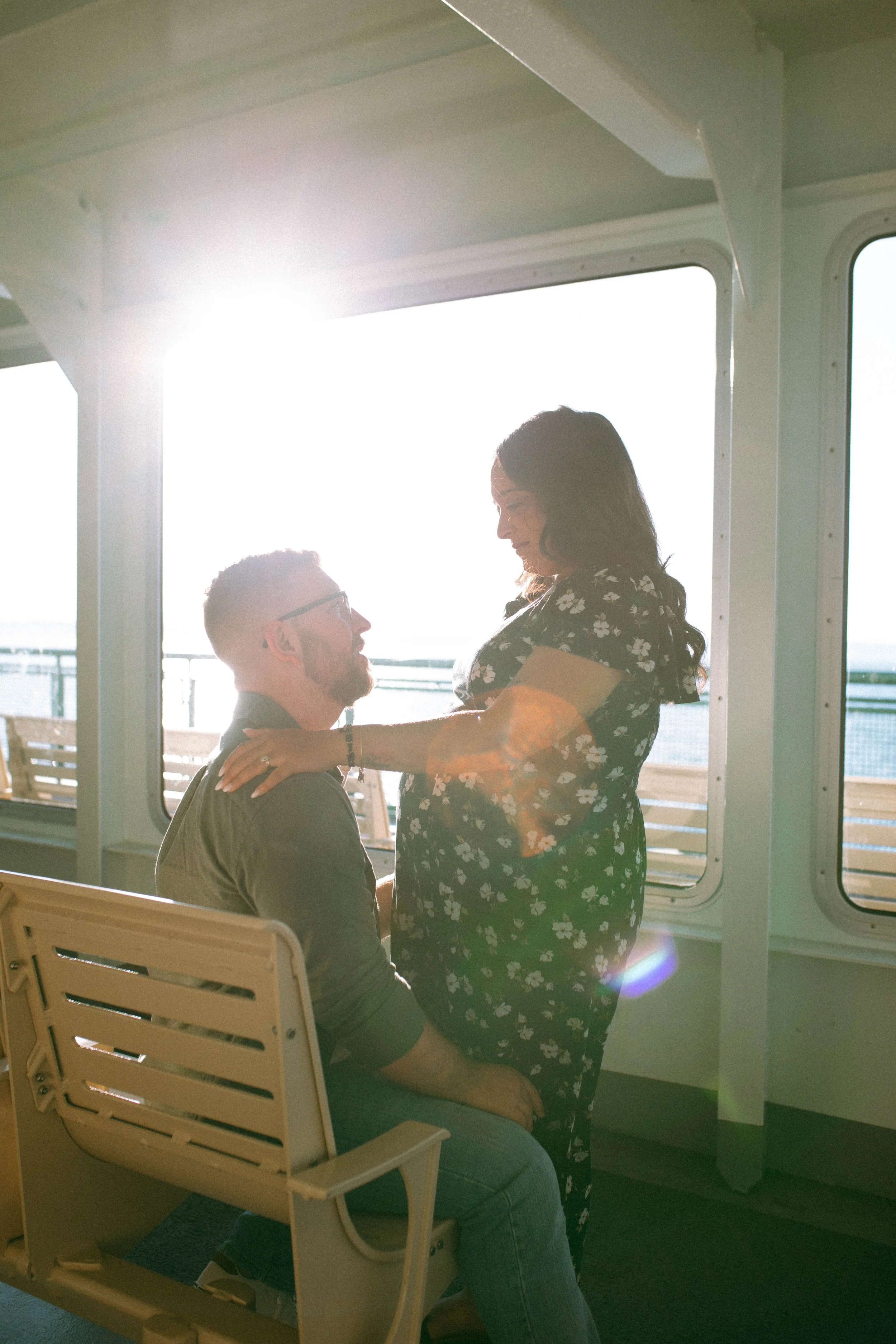 A couple sharing a romantic moment on a cruise ship, with the woman standing and the man sitting, sunlight coming through the windows.