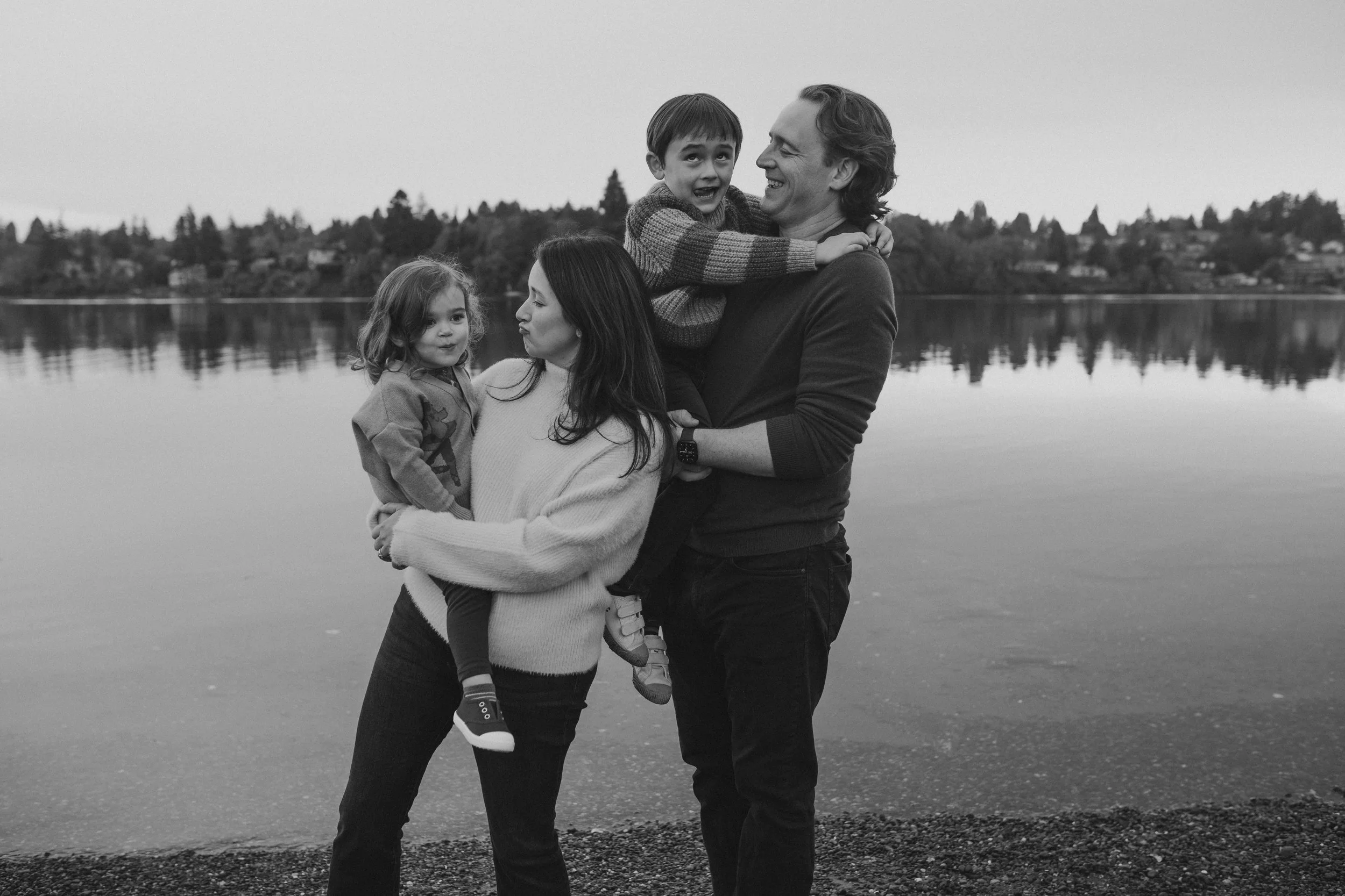 A family of four stands by a lake, embracing and smiling together on a cloudy day.