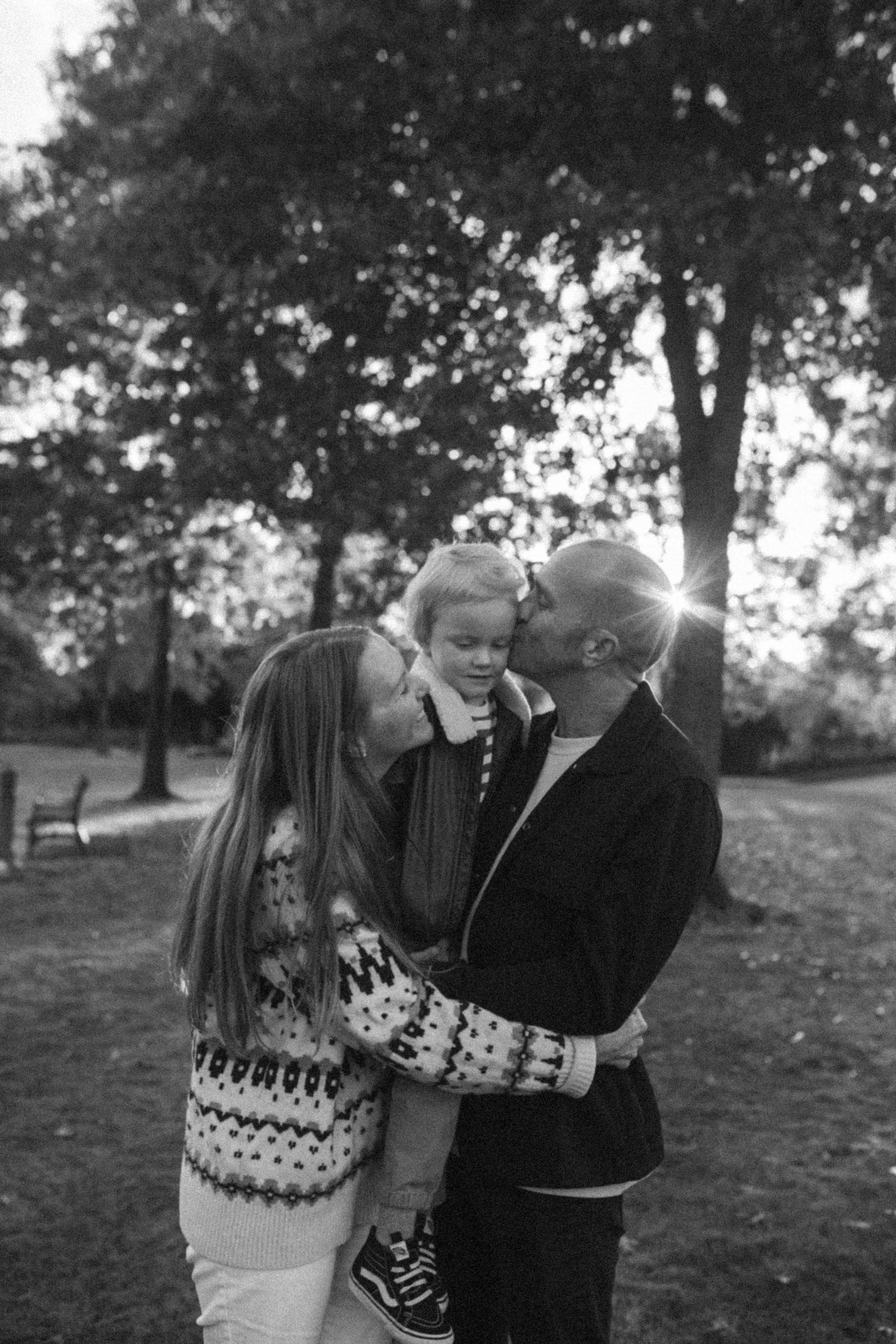 A family of three, a mother, father, and young child, sharing a kiss in a park with trees and benches in the background during what appears to be late afternoon or early evening.