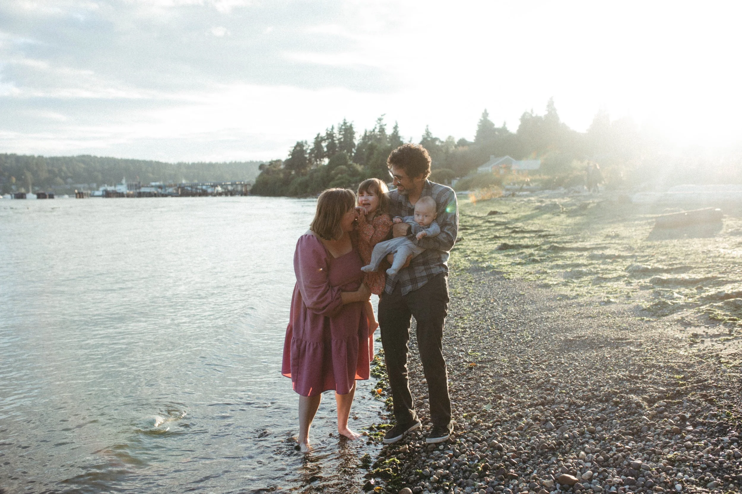 A family enjoying time at a lakeside beach during sunset. An adult woman, an adult man, and two young children are present, with the woman and man holding the children and smiling.