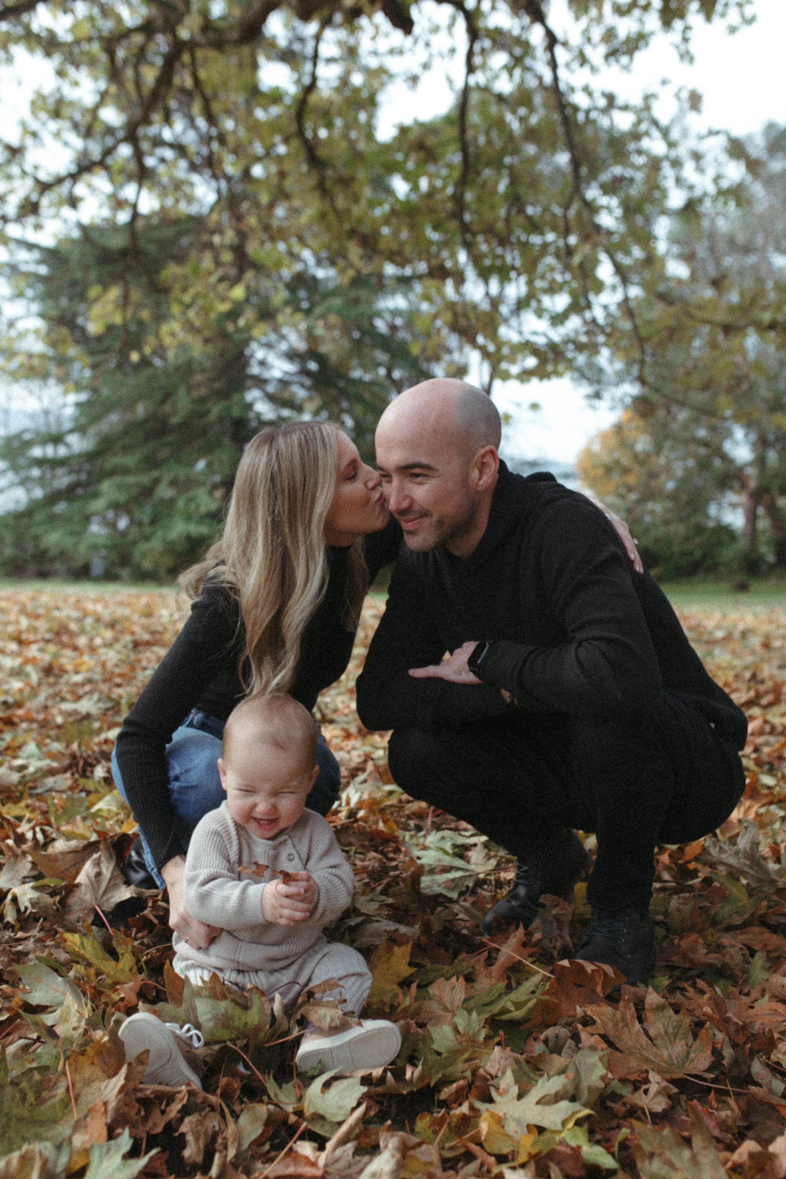 A happy family of three playing outdoors in a park covered with colorful autumn leaves. The mother, father, and their baby are smiling and enjoying a fall day.