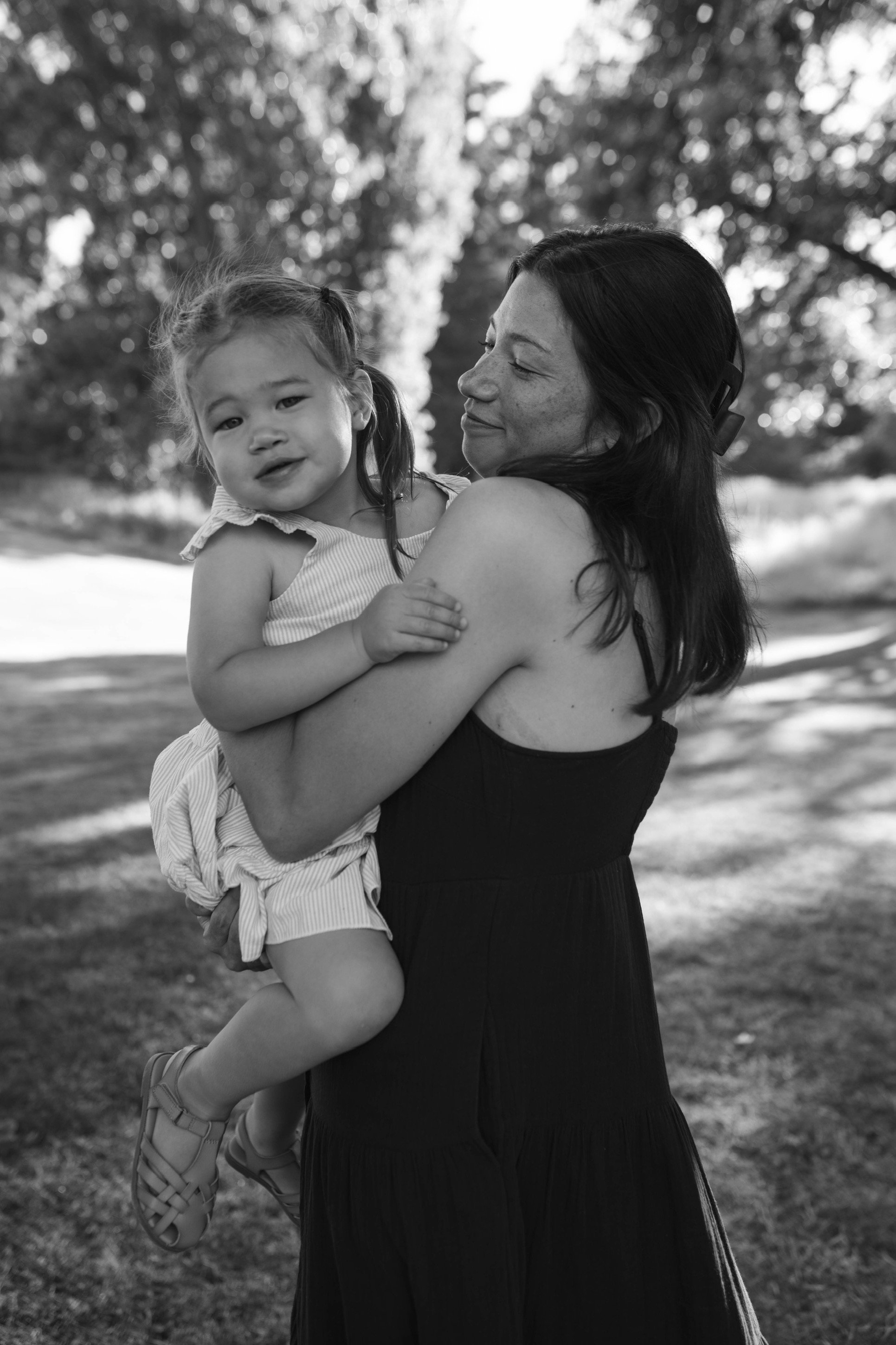 A woman holding a young girl in her arms outdoors in a park, with trees in the background, captured in black and white.