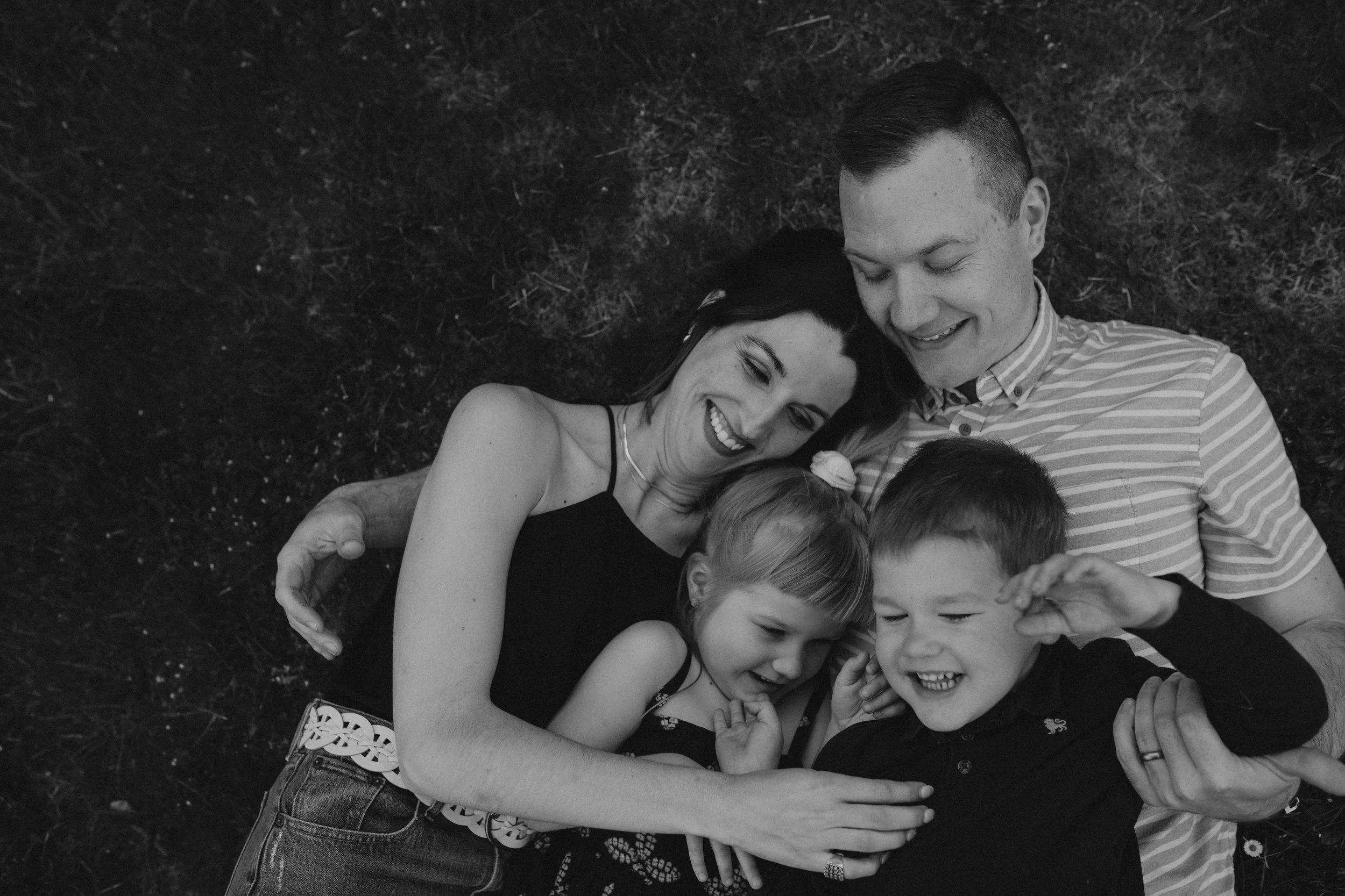 A family of four lying on grass, smiling and laughing together, with the mother and father holding their two children.