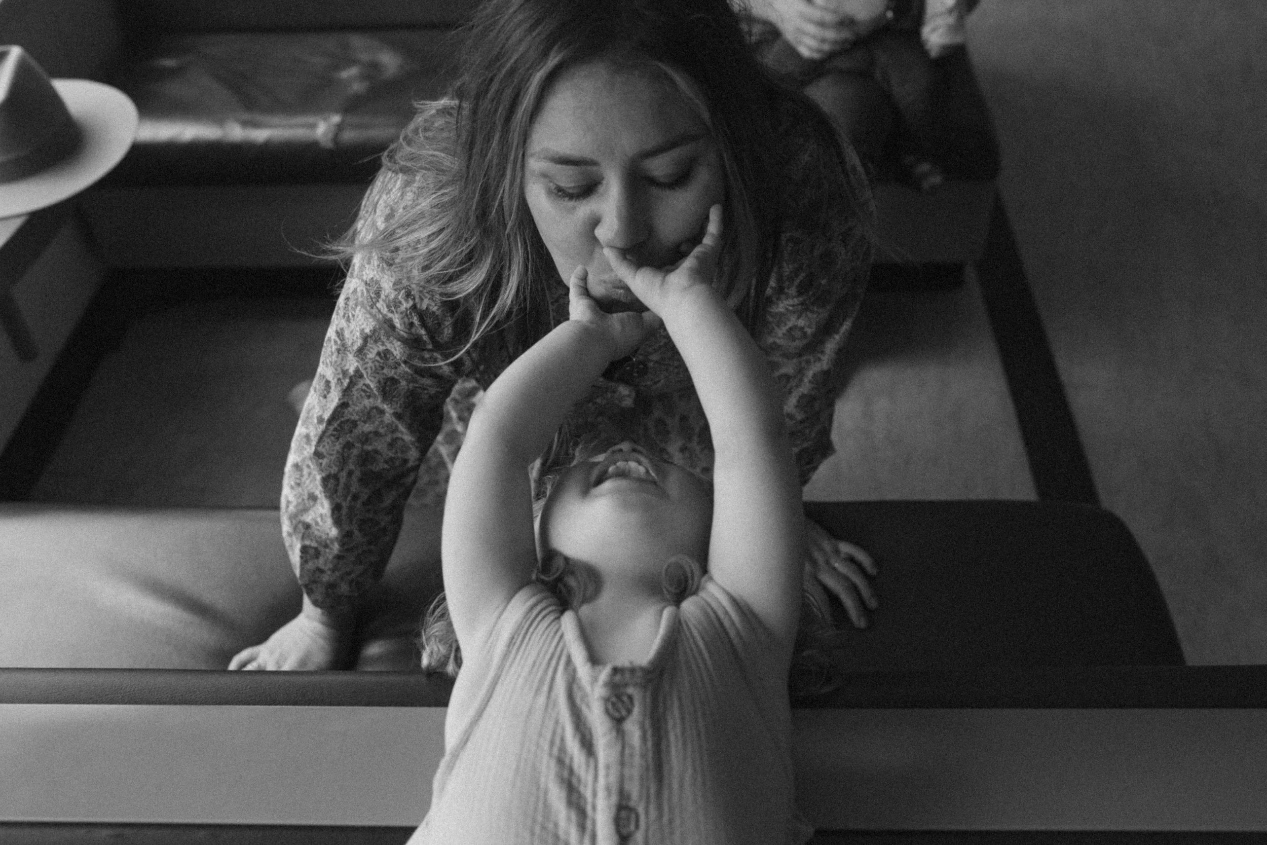 A black-and-white photo of a woman leaning over a child, who is lying on a table or counter with arms raised and mouth open, playfully touching her nose with her finger.