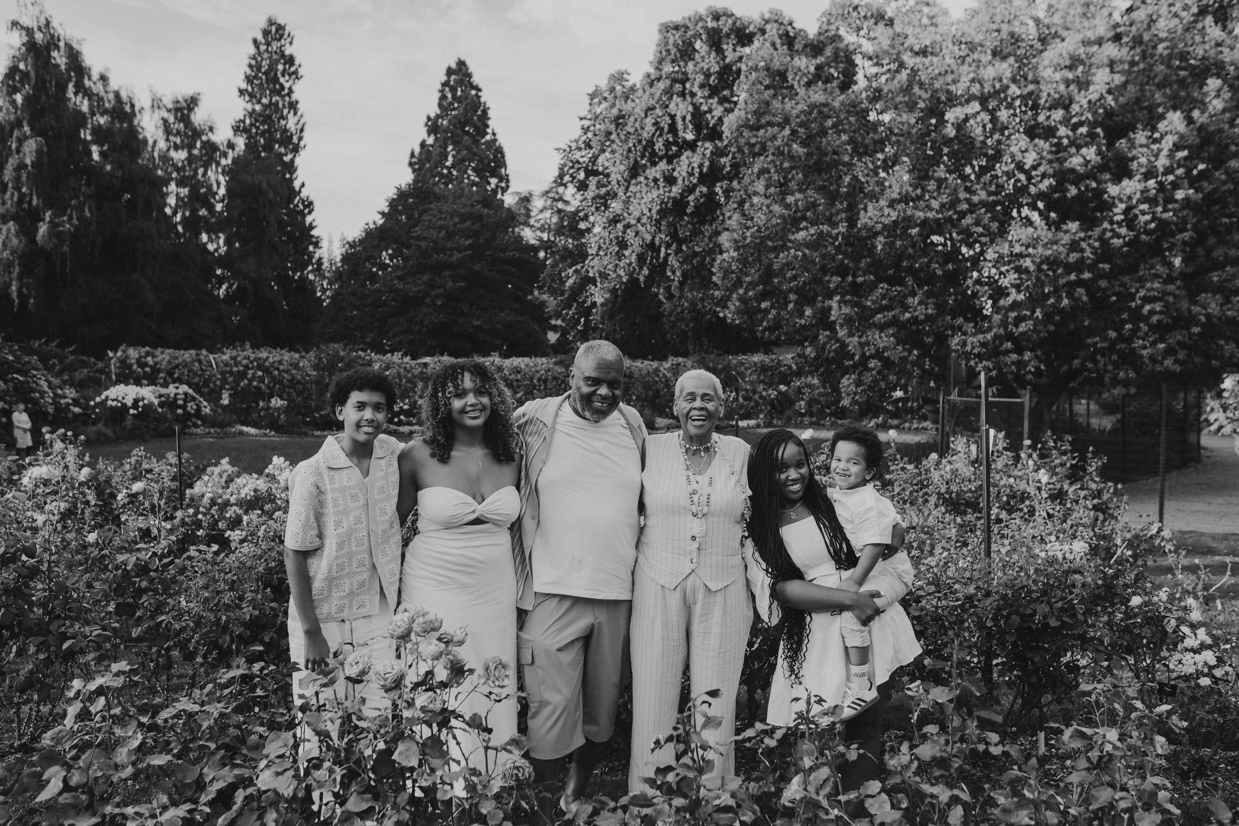 A family of six standing together in a garden with flowers and trees, smiling and posing for the photo.