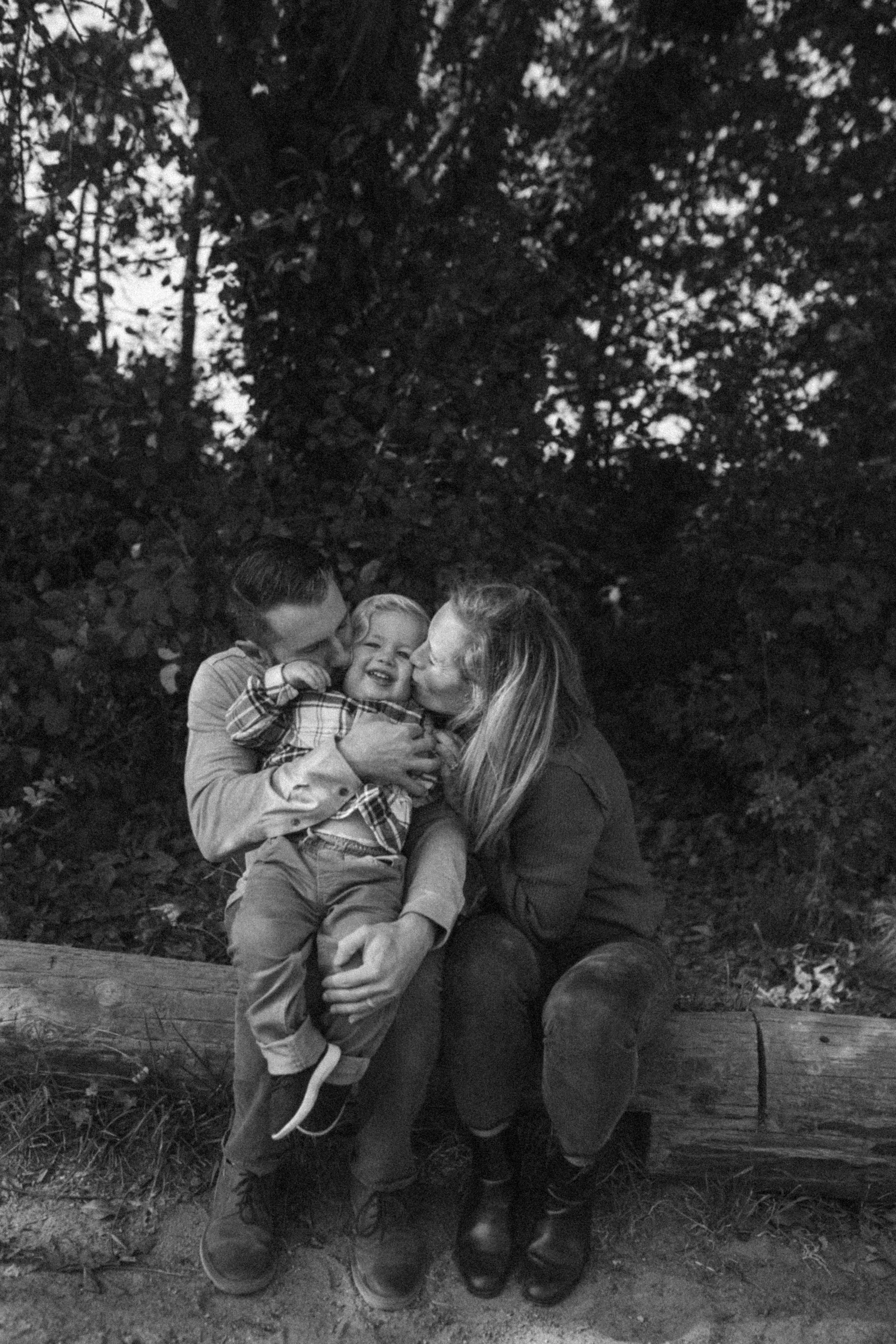A black and white photo of a family sitting on a log outdoors. A man and woman are kissing a young boy in the cheeks, who is smiling happily.