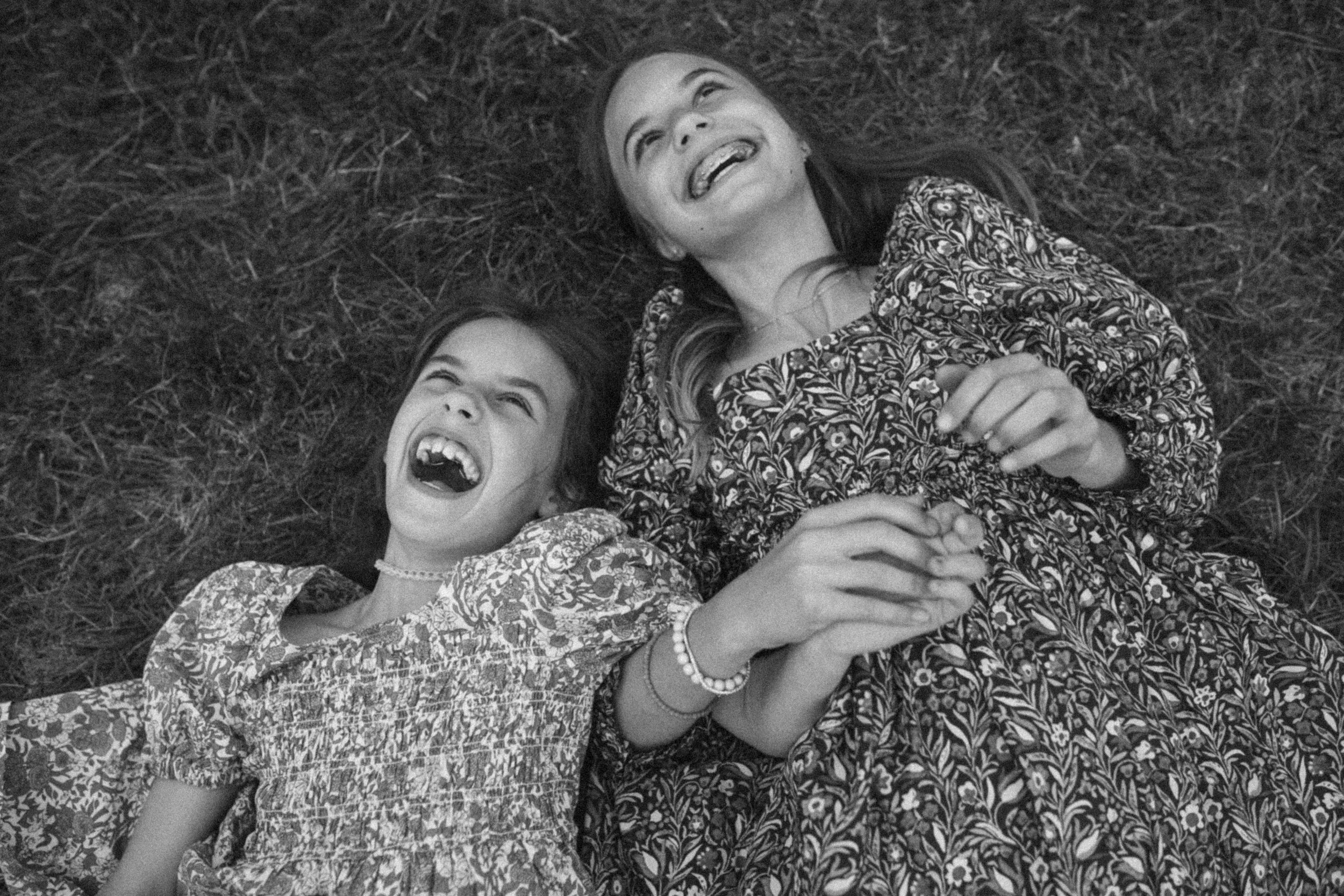 A woman and a young girl lying on the grass, both laughing and holding hands.