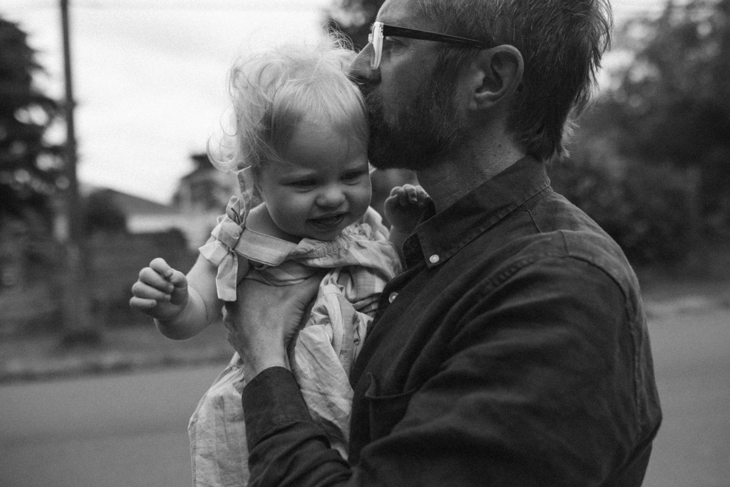 A man with glasses and a beard holding a young girl, who appears to be upset or crying, in an outdoor setting with trees and a house in the background.