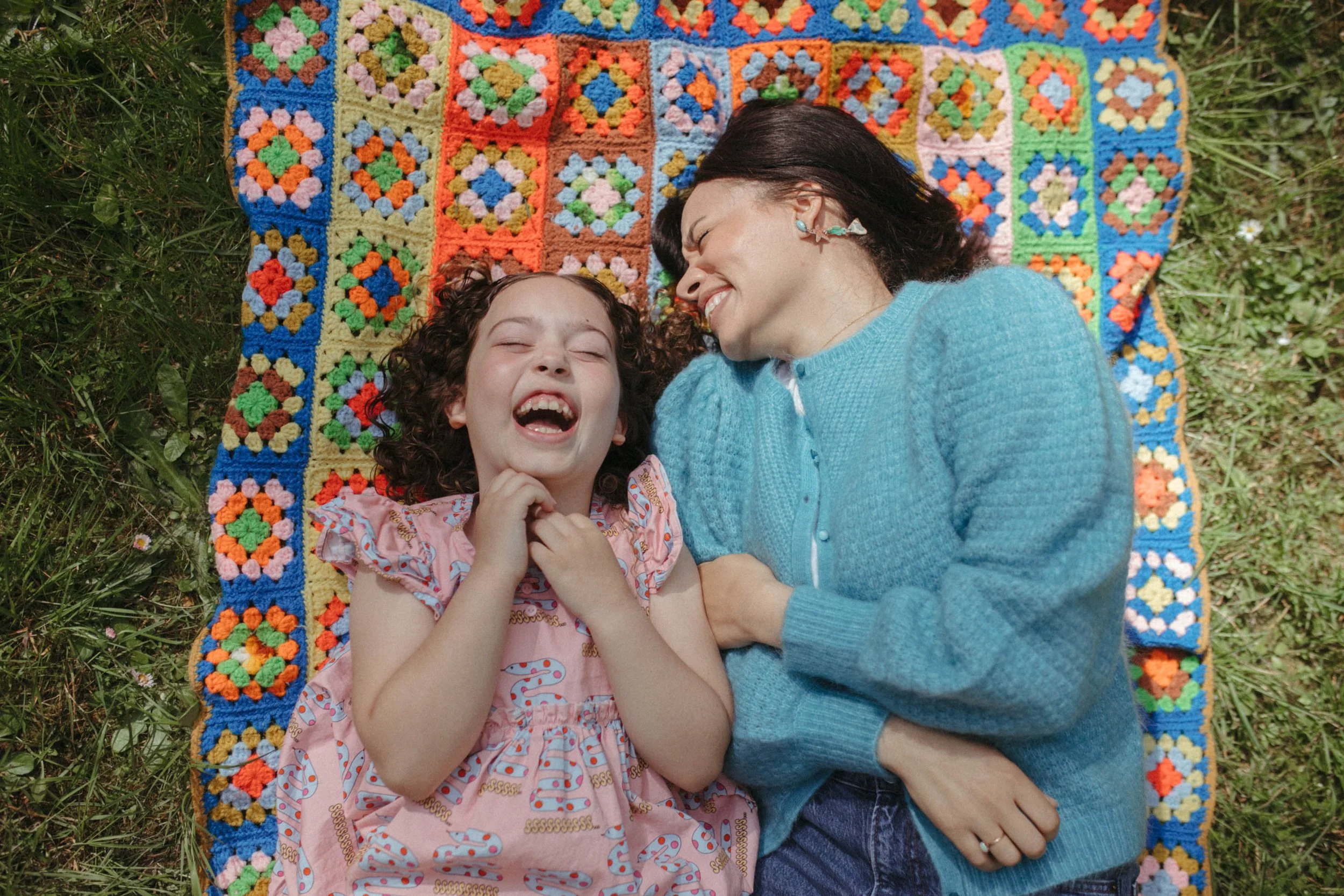 A woman and a young girl lying on a colorful crochet blanket outdoors, smiling and laughing together.