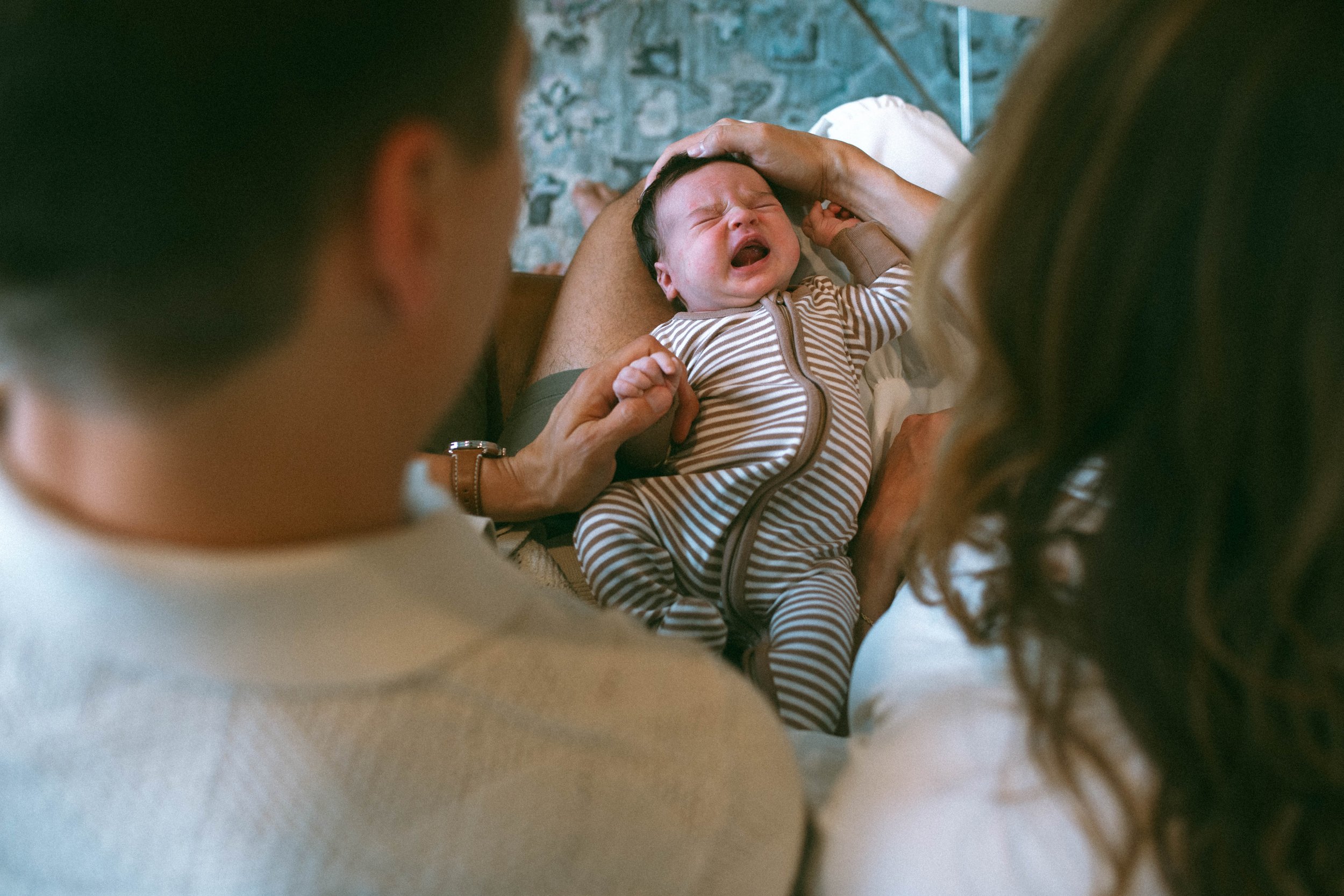 A crying baby in a striped onesie being held by parents in a cozy room with a brick wall background.