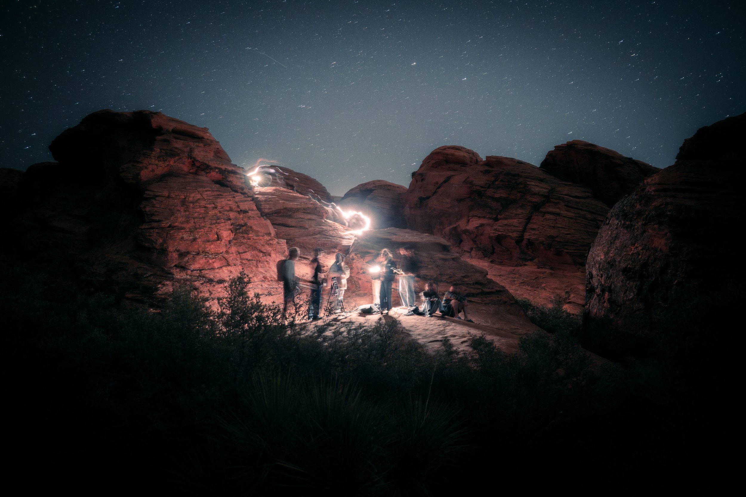 Nighttime desert scene with large red rock formations under a starry sky. A group of people with flashlights are gathered around a light source in the center of the scene.