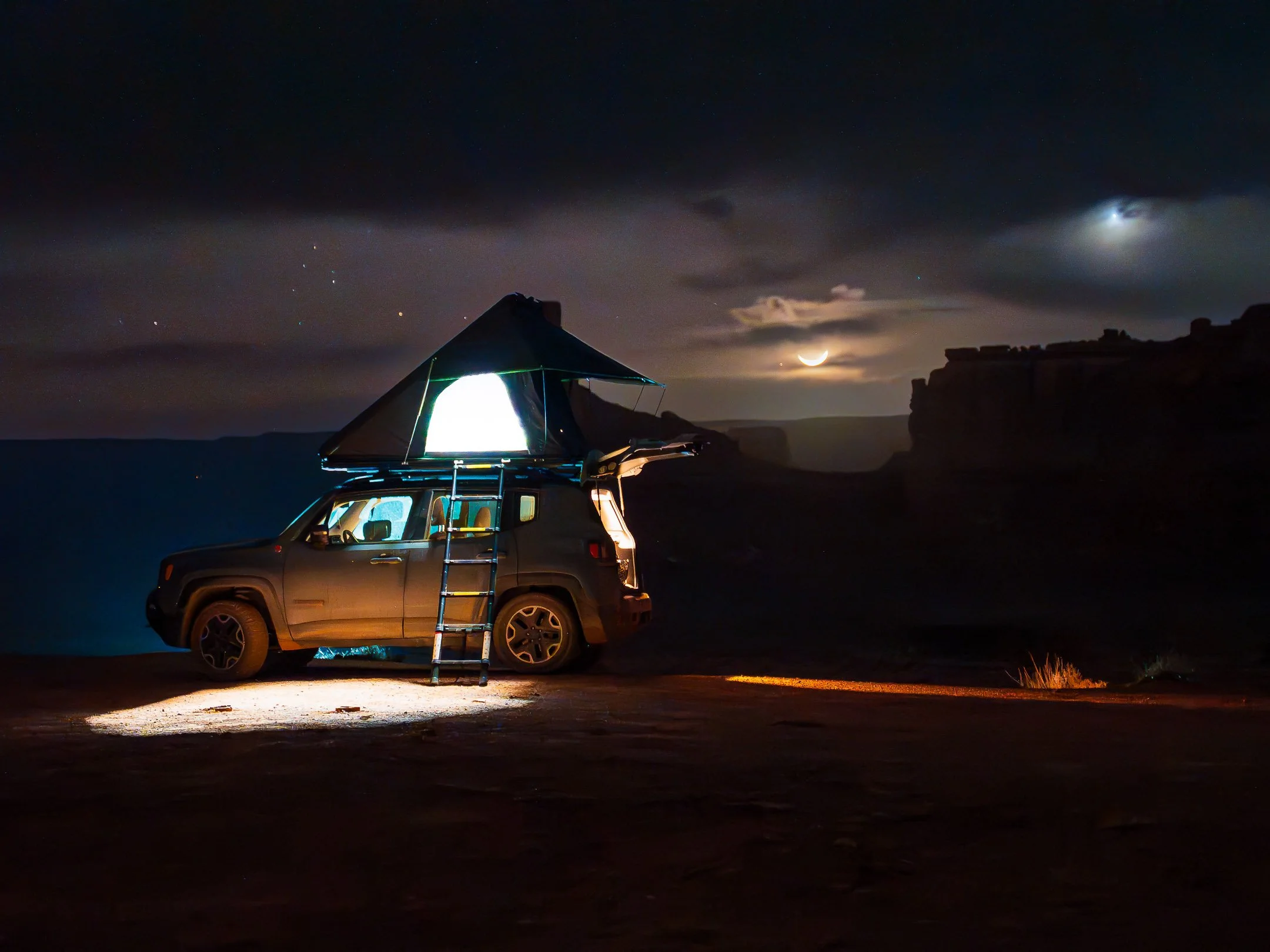 Nighttime scene of a vehicle with a rooftop tent set up, illuminated from inside, parked on a trail with a ladder leading up to the tent. The sky is dark with clouds, visible stars, a crescent moon, and a bright planet or star in the background.