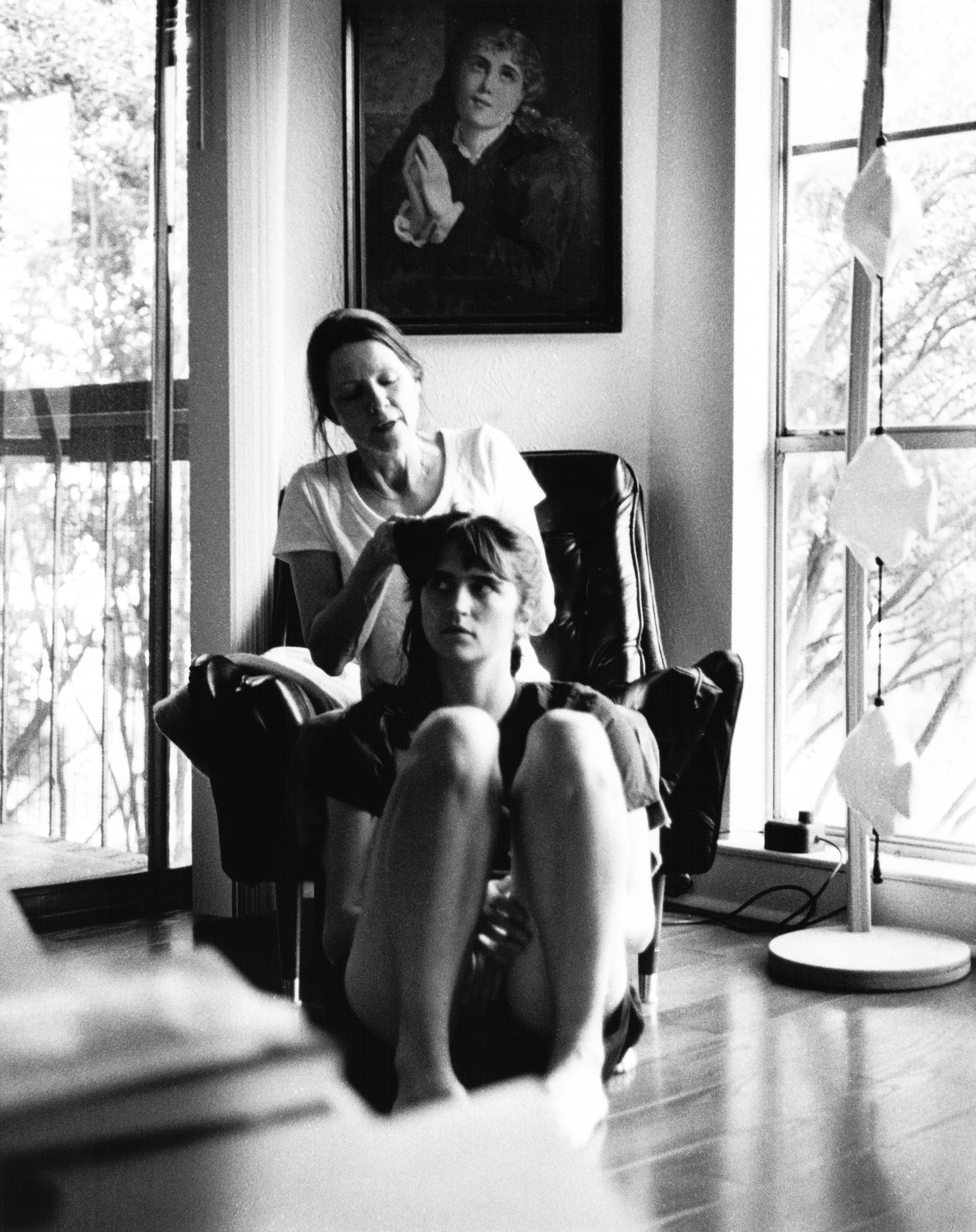 A woman is styling a young woman's hair in a room with a large window. A portrait hangs on the wall behind them, and a small lamp is on the floor to the right.