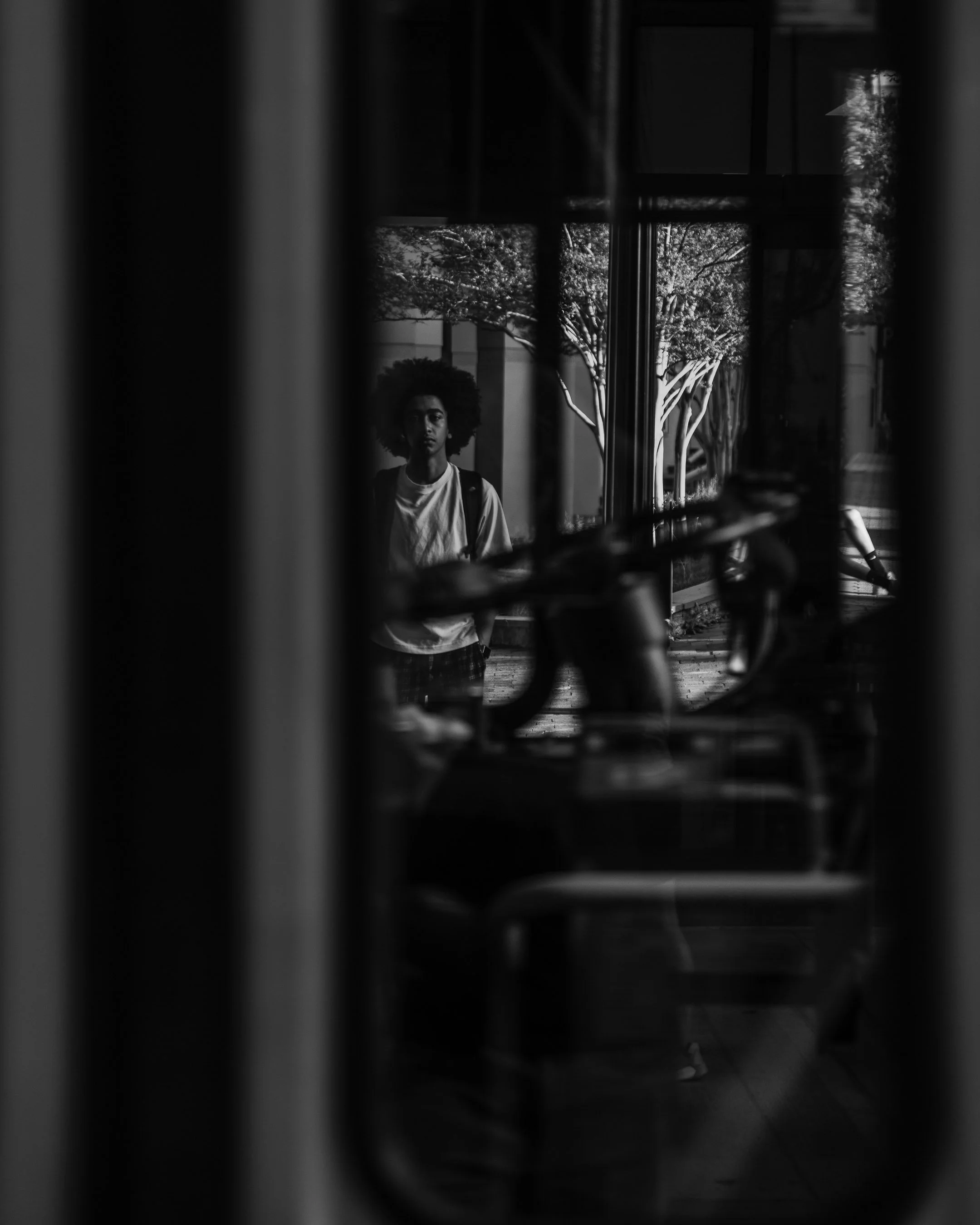 Black and white photo of a person with an afro hairstyle standing on the street, looking through a bus window.