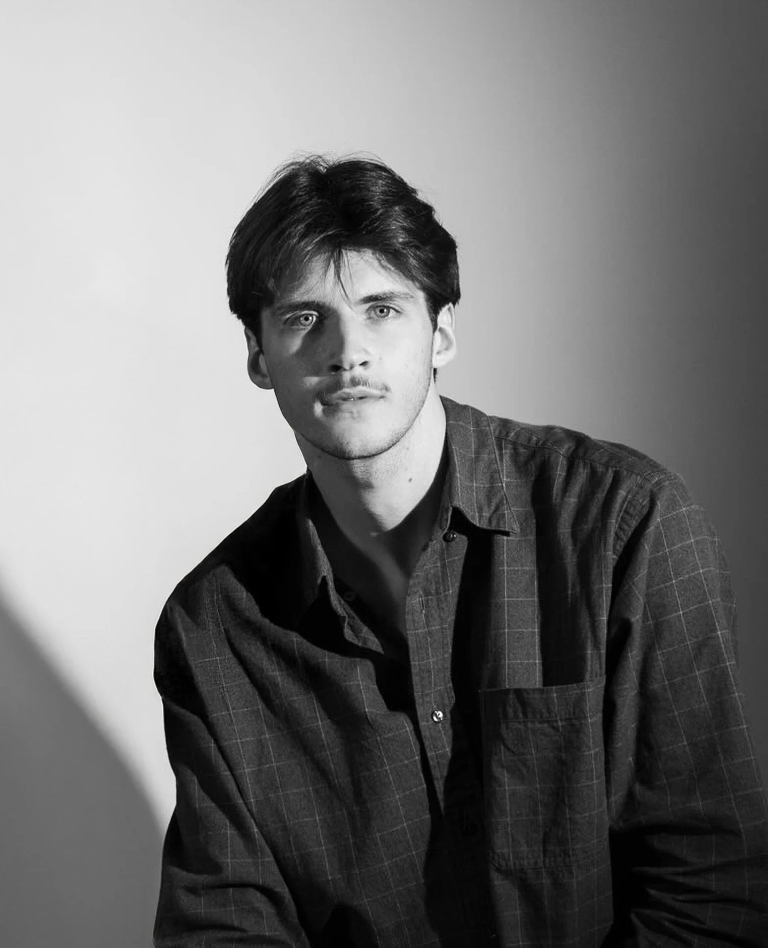 A black-and-white portrait of a young man with dark hair, wearing a plaid shirt, looking into the camera with a serious expression.