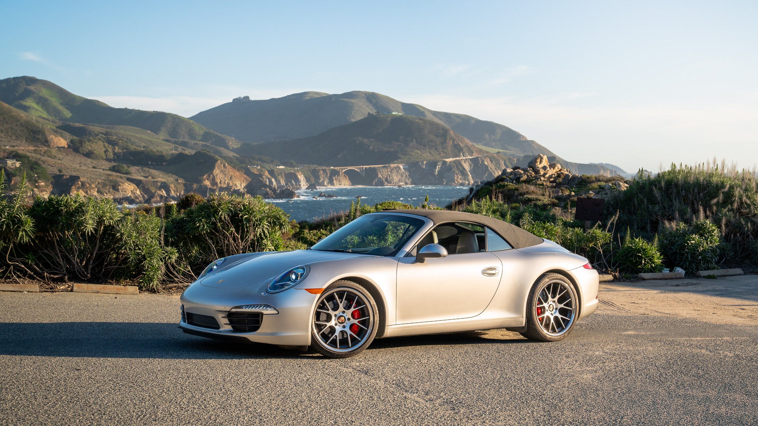 A silver Porsche convertible parked on the side of a scenic coastal road with green hills, rocks, and ocean in the background.