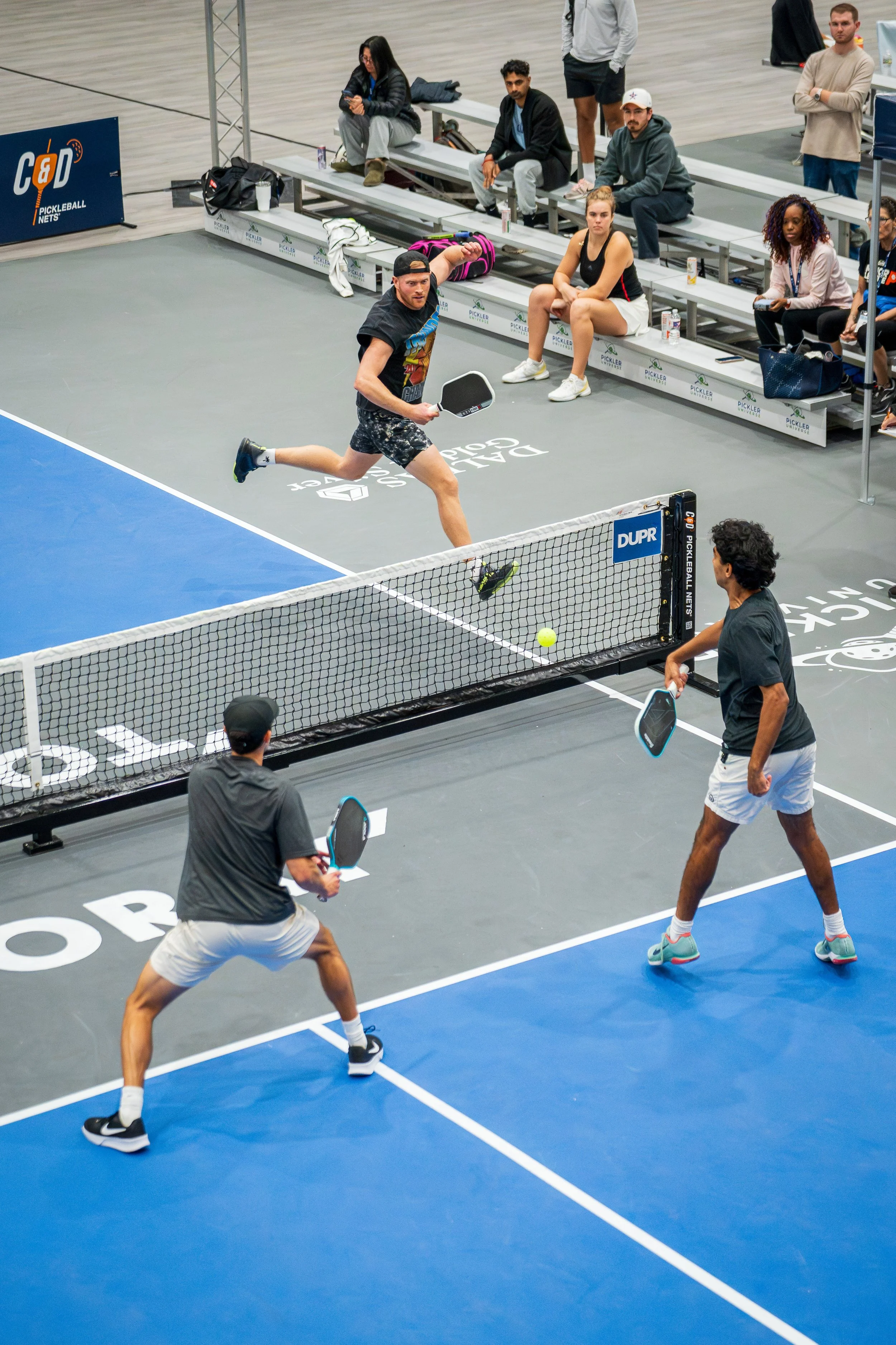 Two men playing pickleball on an indoor court while a third man hits a ball over the net. Spectators sit on a bench along the wall, watching the game.
