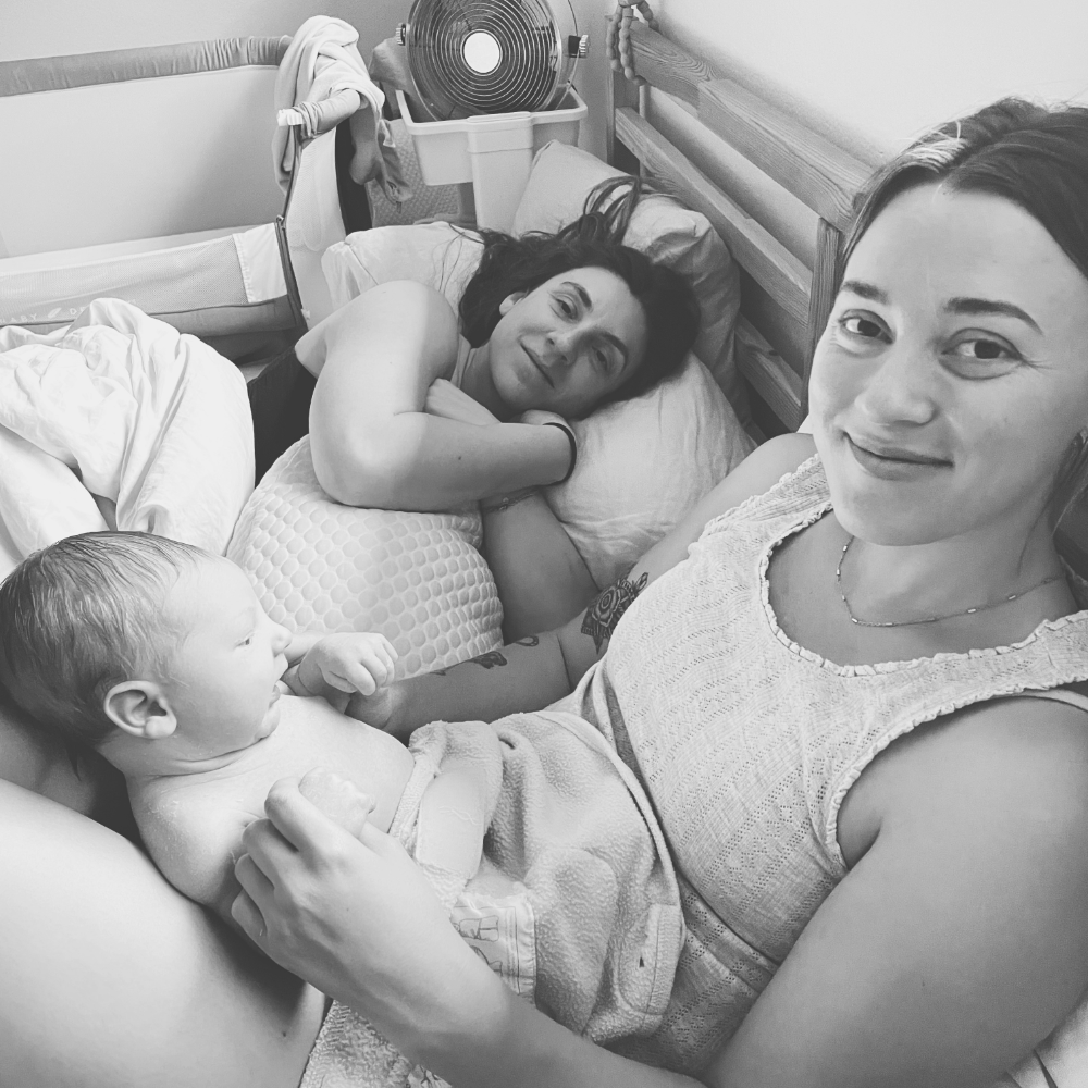 4 The Moms Southern California Postpartum Doula testimonial photo, black and white, of a woman holding a baby on her legs, with another woman lying in bed in the background. The scene is home recovery setting for the 4th trimester.