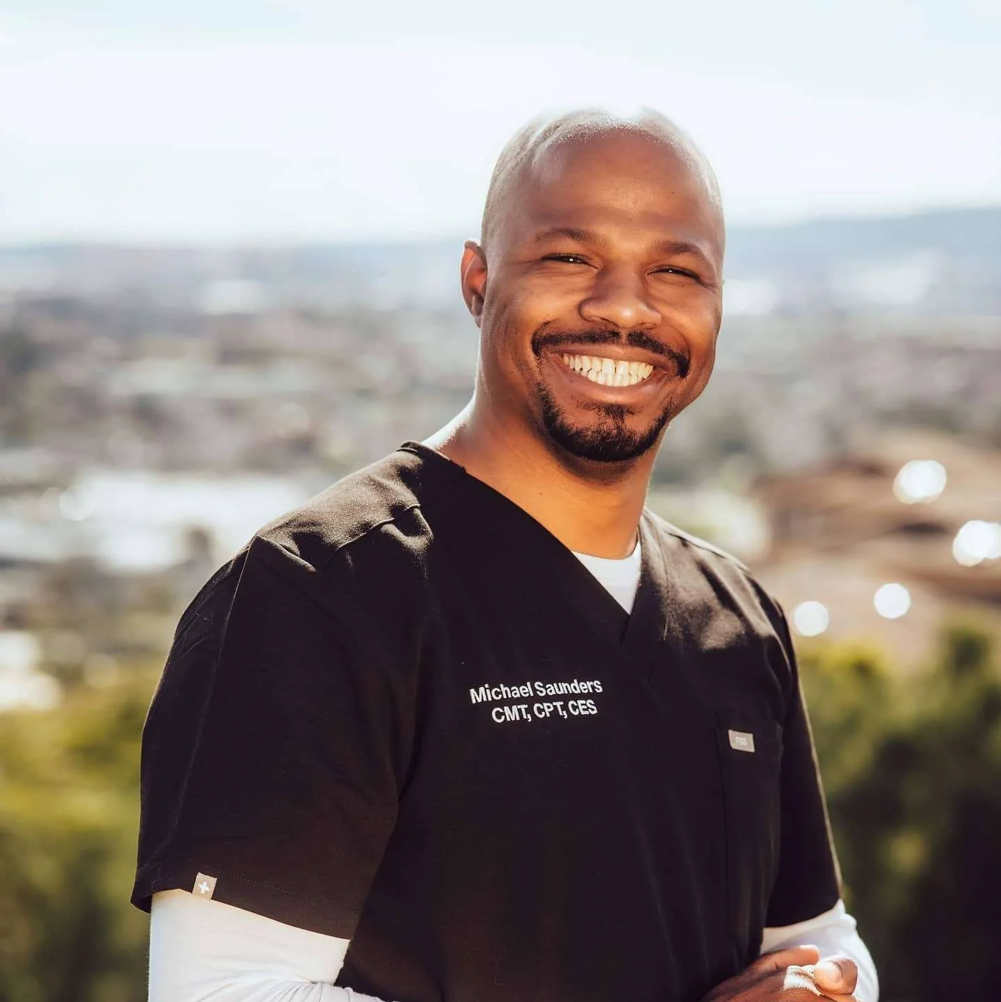 A smiling man in a black medical uniform outdoors with a blurred cityscape background.