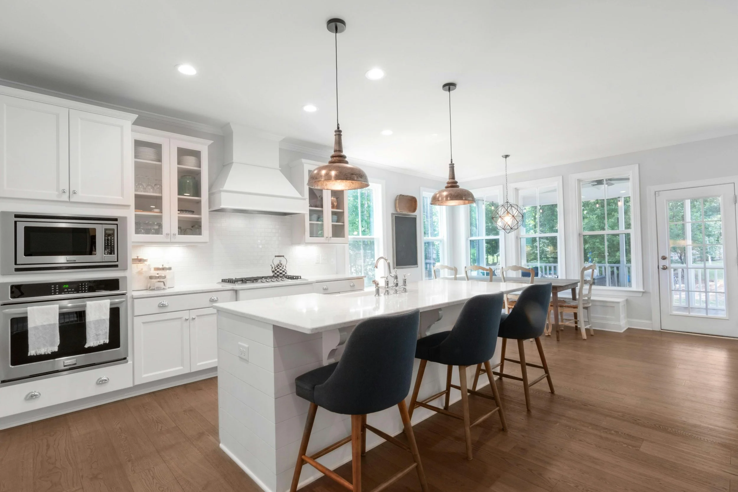 Modern white kitchen with an island, black bar stools, copper pendant lights, white cabinets, and a dining area with large windows and a door to the outside.
