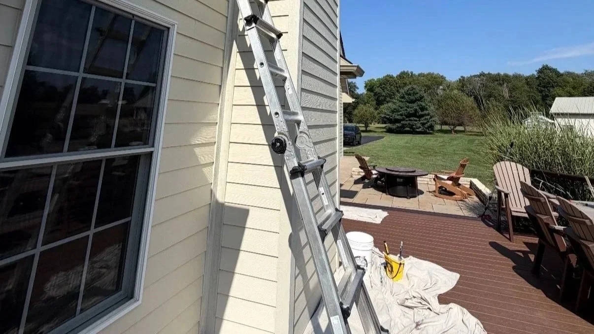 Extension ladder leaning against a house beside a window, with a backyard patio, outdoor furniture, and painting supplies.