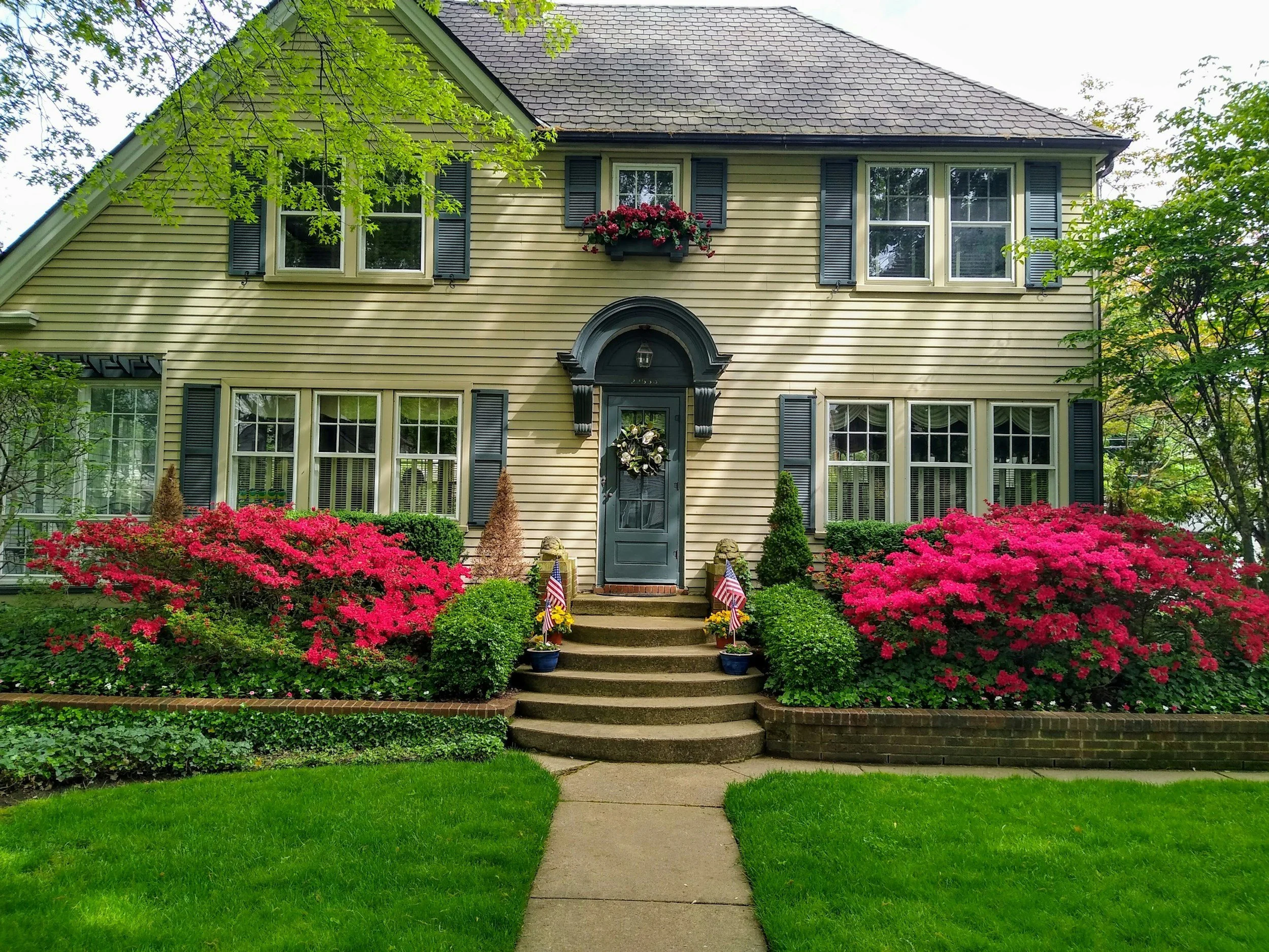 A two-story house with beige siding, black shutters, and a gray door with a floral wreath. The house has large windows and a front yard with vibrant pink and red flowering bushes, green shrubs, and a green lawn. Two small American flags are placed on each side of the front steps.