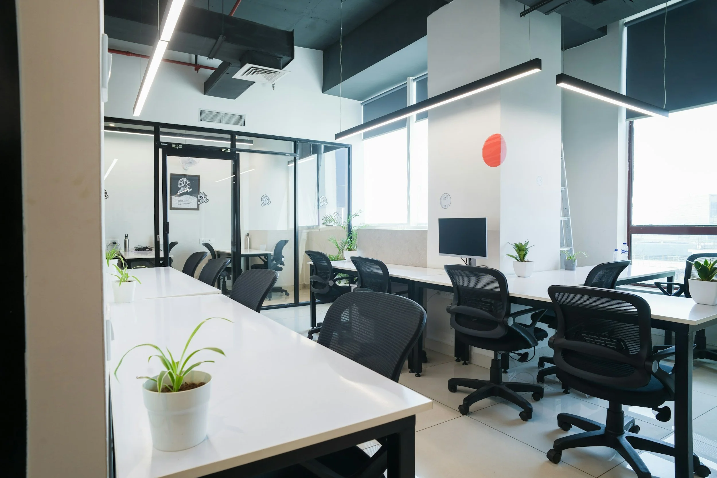 Modern office with white desks, black ergonomic chairs, potted plants, and large windows letting in natural light.
