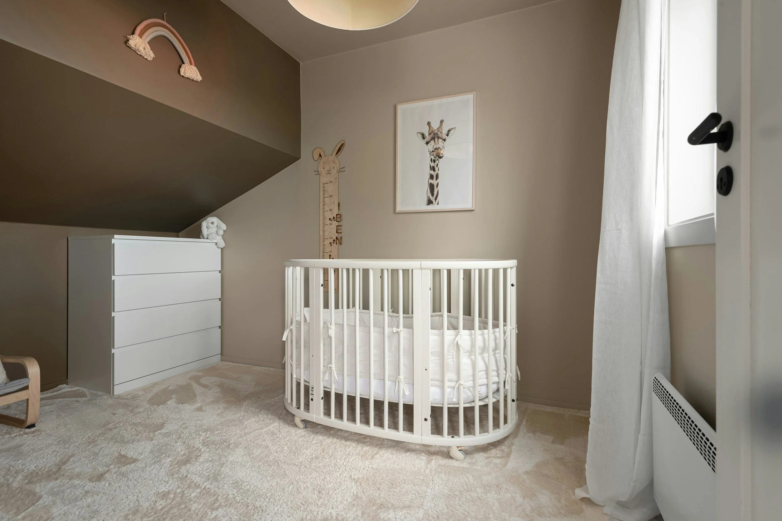 Nursery room with a white crib, a funny height measurement chart with a rabbit, framed giraffe picture, white dresser, and rainbow wall decor, with a window covered by a white curtain.