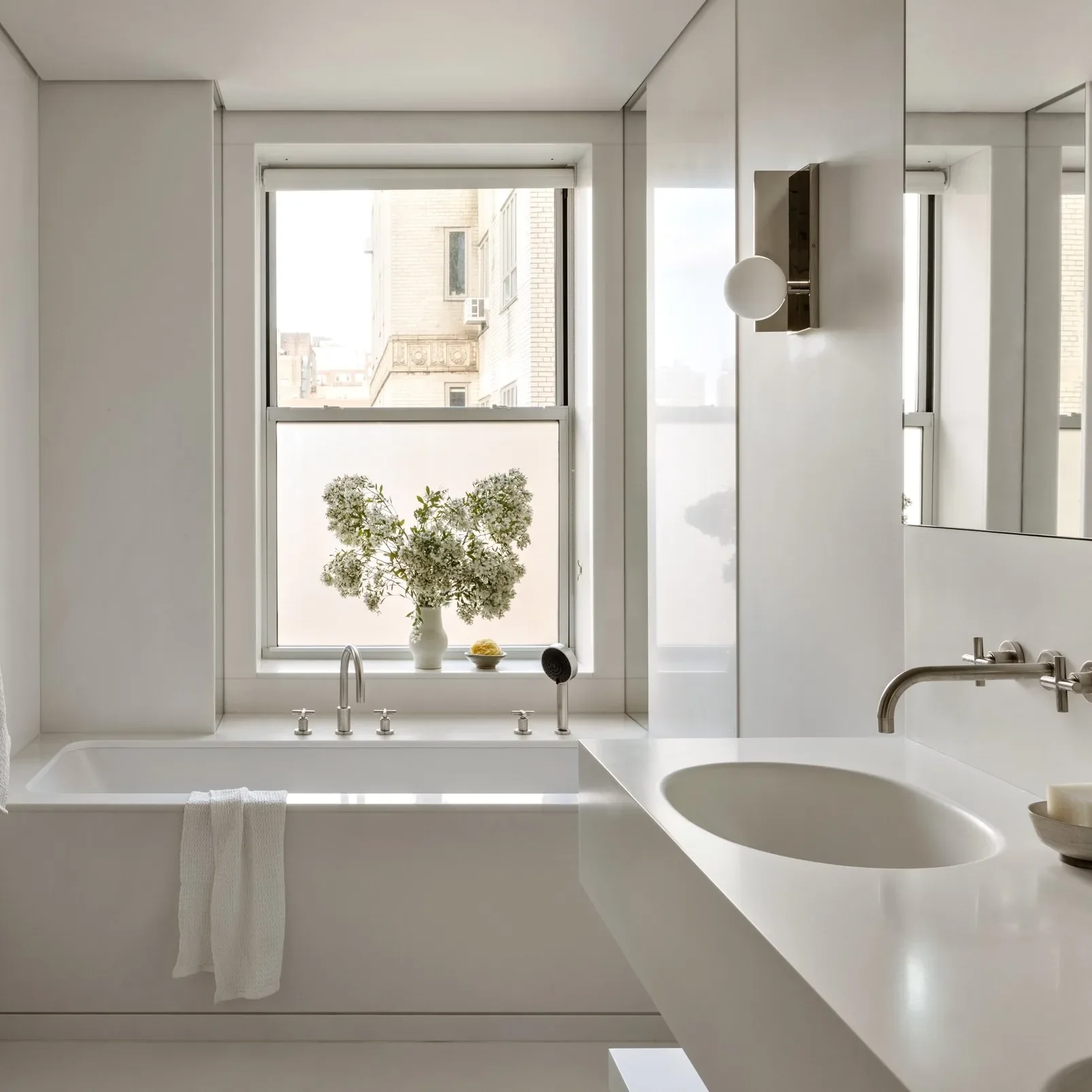 Bright modern bathroom with large window, white countertop, sink, and a tub with a towel hanging on the edge. Decor includes a white vase with flowers and a yellow decorative item.