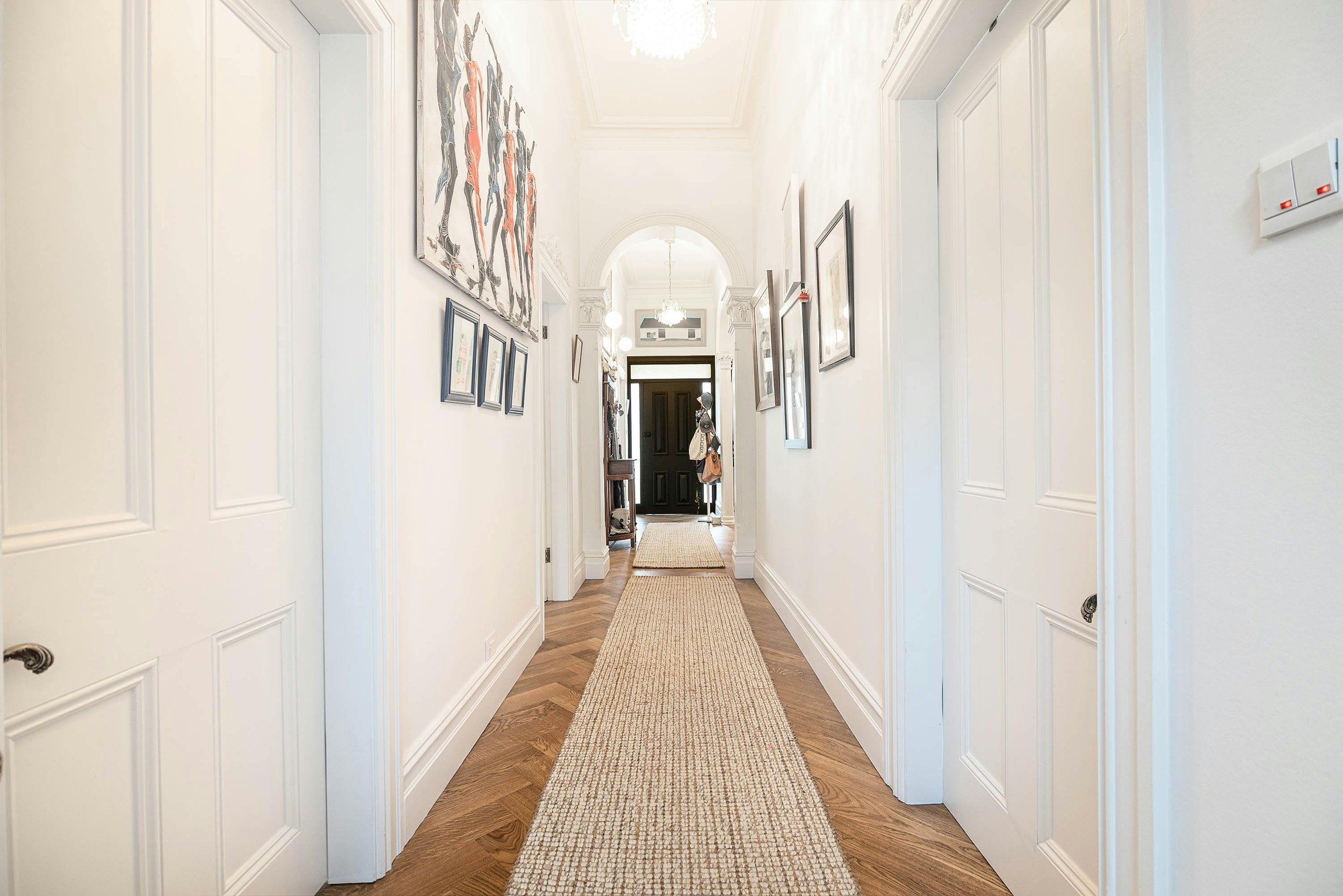 Bright hallway with framed artwork on white walls, wood flooring, beige runner rug, leading to a black front door with a coat rack inside.