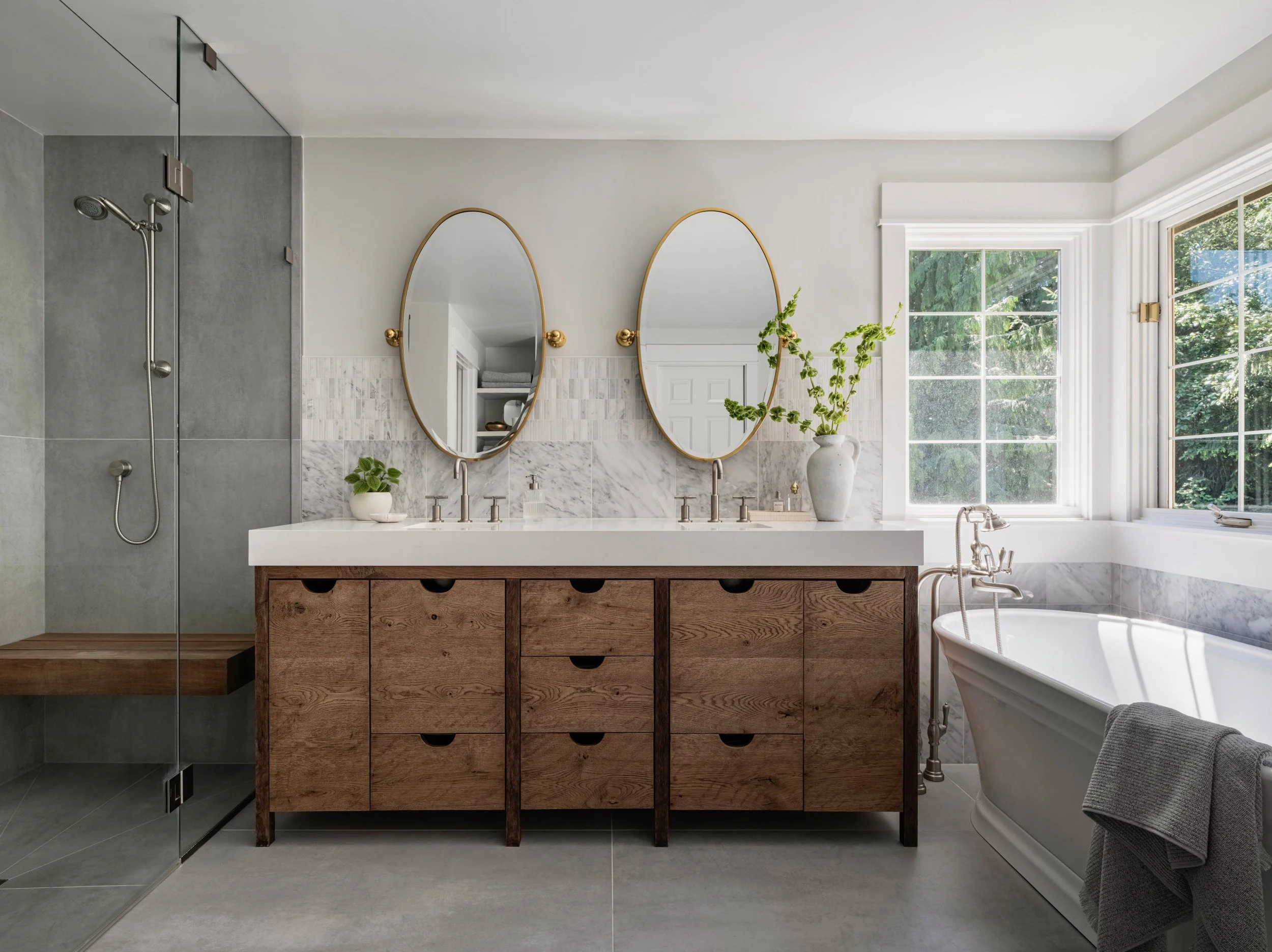 Modern bathroom featuring a large wooden vanity with two sinks, two oval mirrors with gold frames, a white marble countertop, a potted plant, a white bathtub, a window with greenery outside, and a glass shower enclosure.