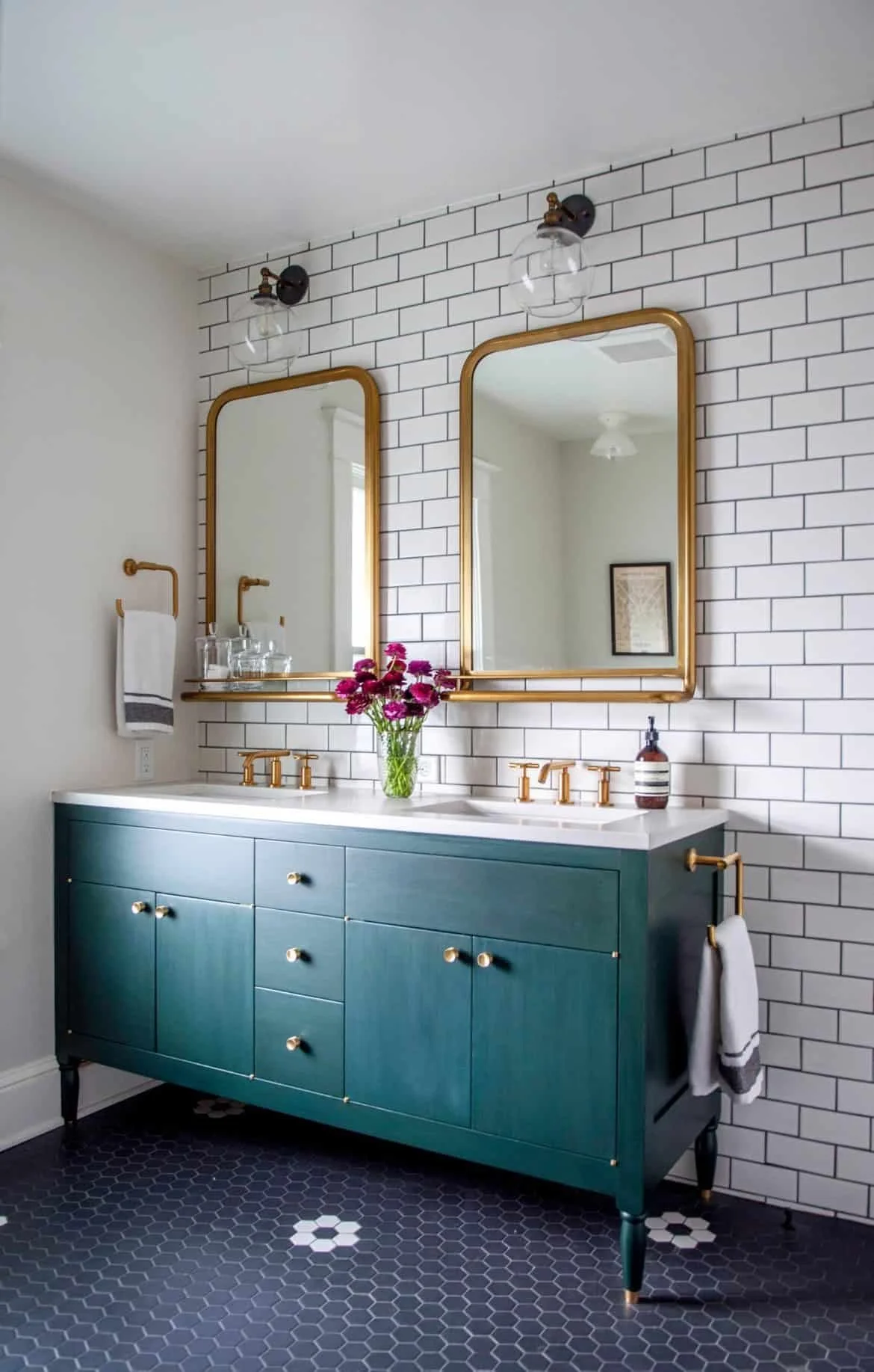 A bathroom with a teal vanity, two large gold-framed mirrors, white subway tile walls, black hexagonal floor tiles, and modern light fixtures. A vase with pink flowers and a soap dispenser are on the vanity. Hand towel rack with a white towel is on the wall.