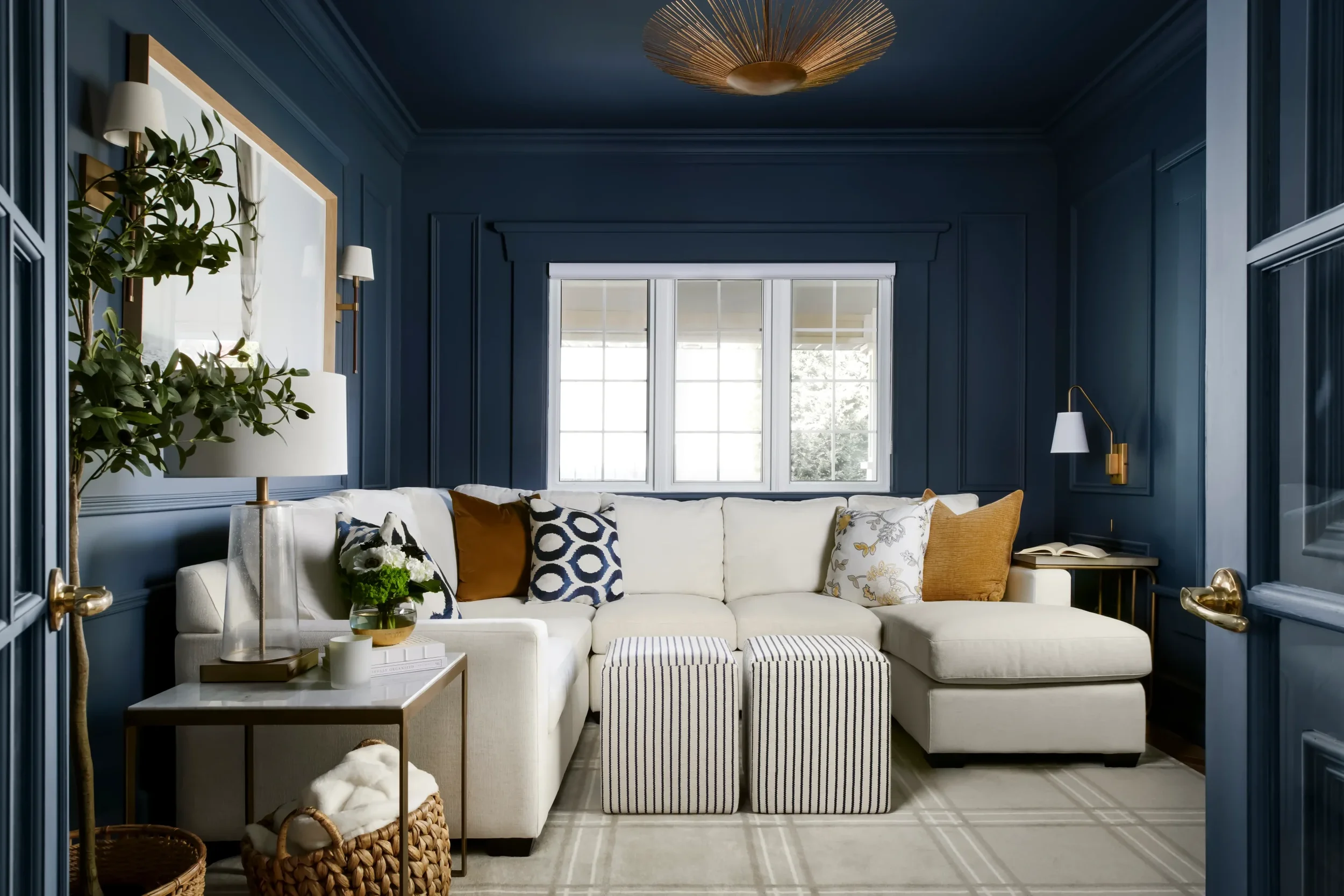 Living room with dark blue walls, white sofa with patterned pillows, a glass side table with books and a flower arrangement, striped ottomans, a ceiling light fixture, and a large window letting in natural light.
