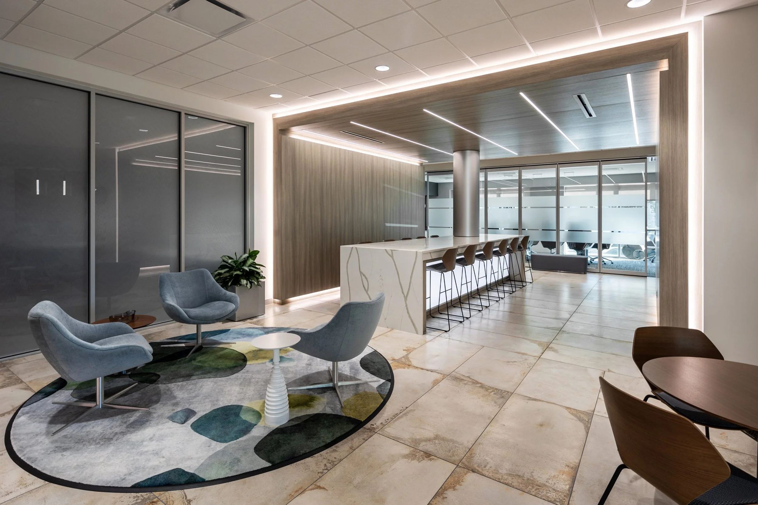 Modern office lounge with blue chairs, patterned rug, marble island bar, and glass conference rooms