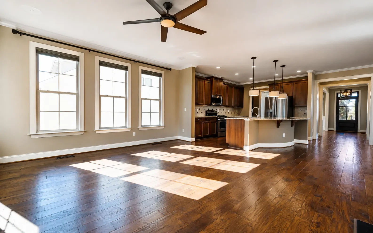 Empty living room with three large windows, hardwood floors, ceiling fan, and an open kitchen with wooden cabinets, a marble island, and pendant lighting.
