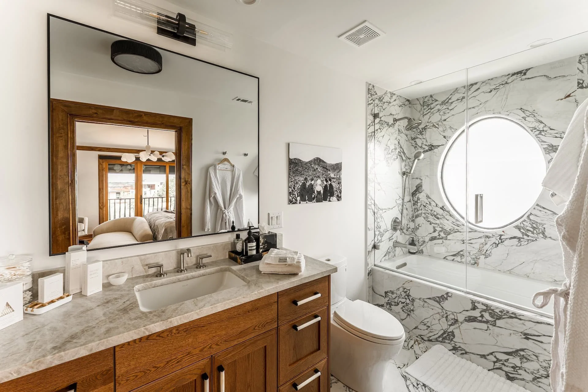 Modern bathroom with marble shower, round window, wooden vanity, medicine cabinet mirror, and artwork on the wall.