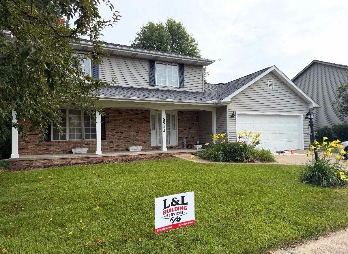 A two-story house with brick and gray siding, a front porch, and a garage, surrounded by a green lawn and some blooming yellow flowers. A sign in the yard advertises L&L Building Services.