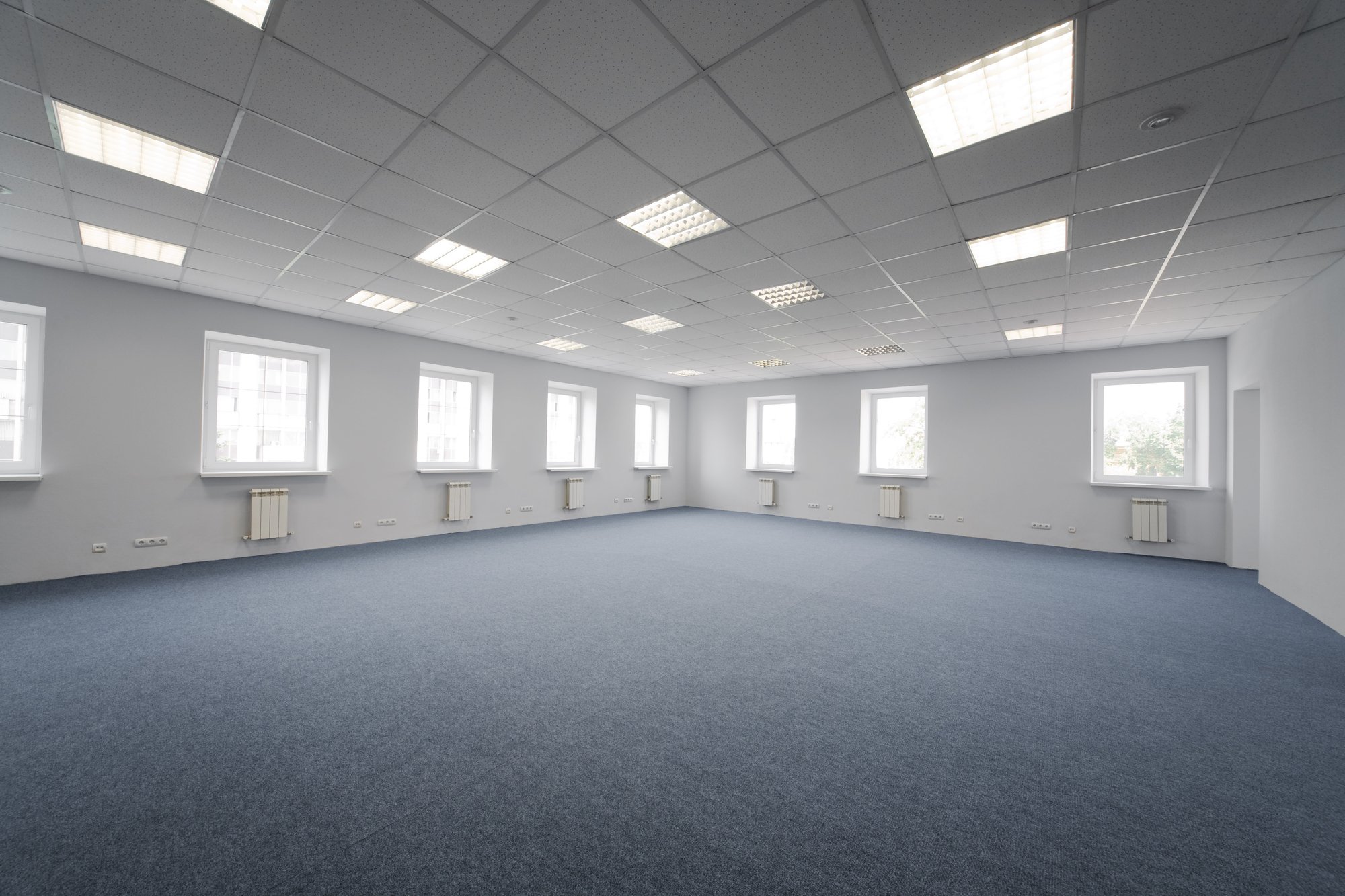 Spacious empty office room with gray carpet, white walls, multiple windows, radiators, and fluorescent ceiling lights