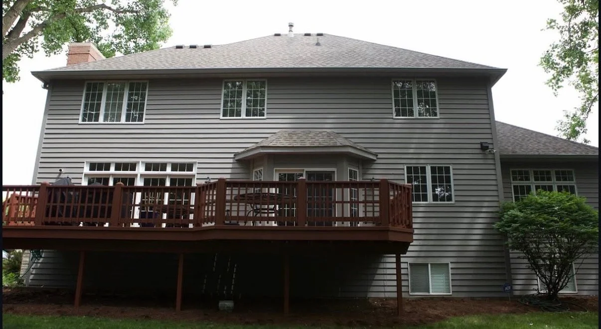 Rear of a two-story house with beige siding, a wooden deck, and multiple windows, surrounded by trees and greenery.