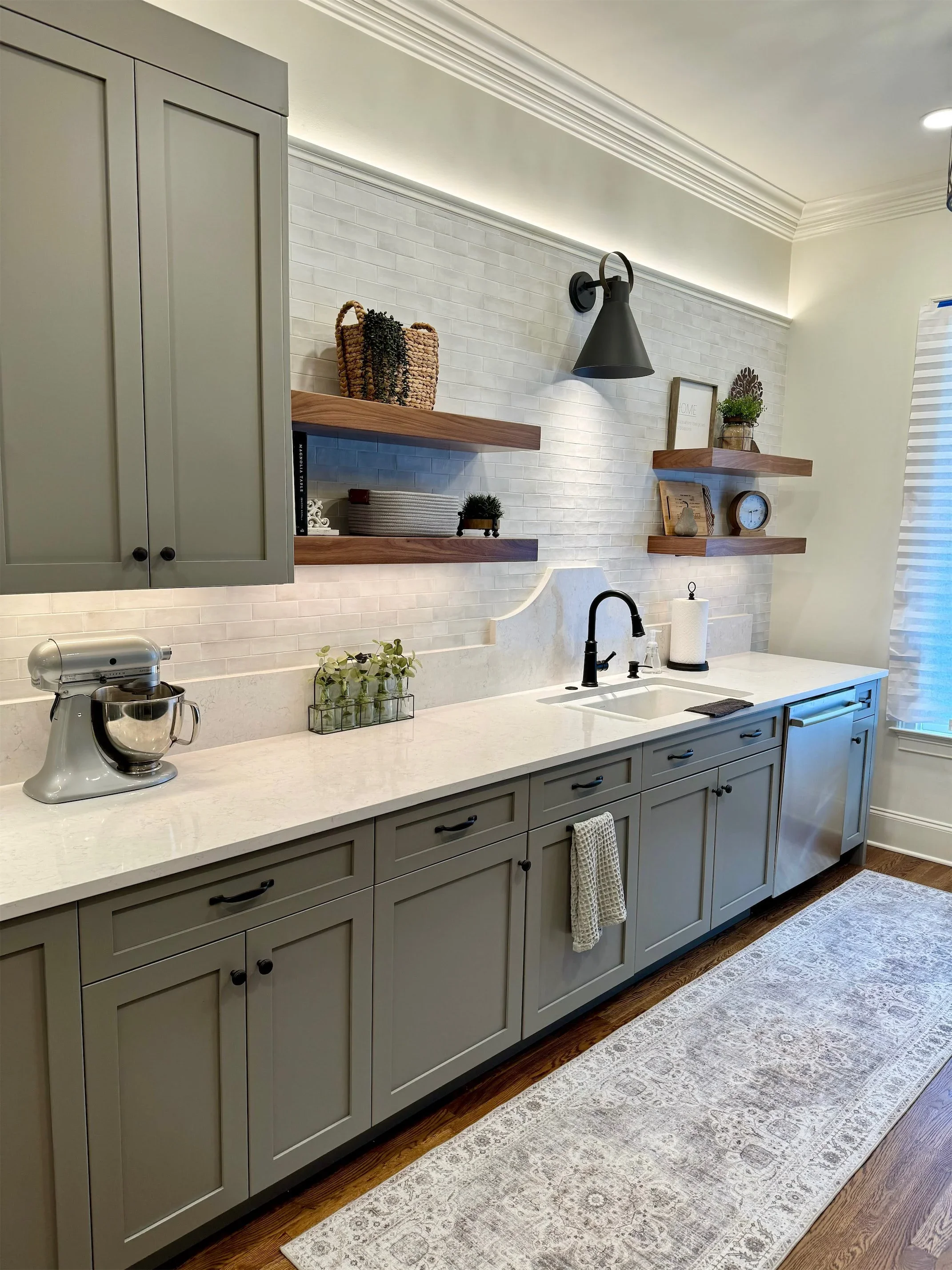 Kitchen with light gray cabinets, white countertop, black faucet, wall-mounted wooden shelves with decor, and a white backsplash wall. Rug on hardwood floor.