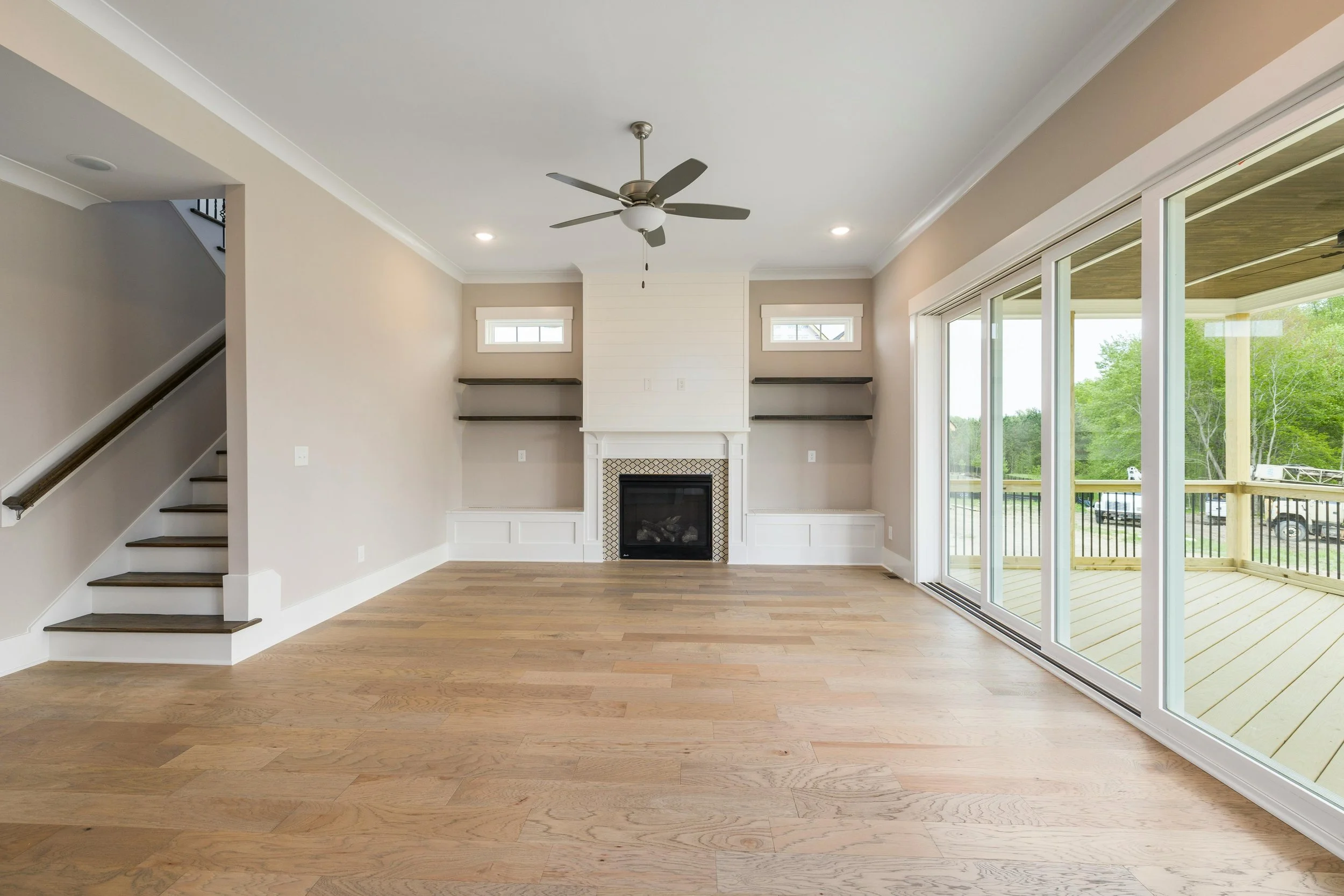 Empty living room with hardwood floors, white walls, a ceiling fan, a fireplace flanked by two small windows, and large sliding glass doors leading to a deck with a view of trees and parked vehicles.