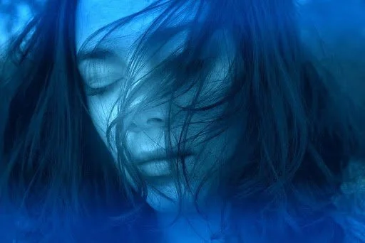 Close-up of a woman with dark hair covering part of her face, with a blue-tinted background.