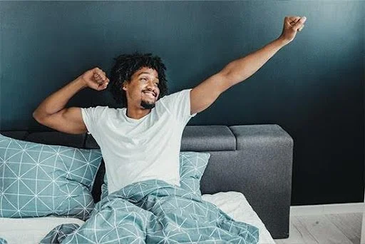 A man smiling and stretching while sitting up in bed with a blue patterned blanket and pillows