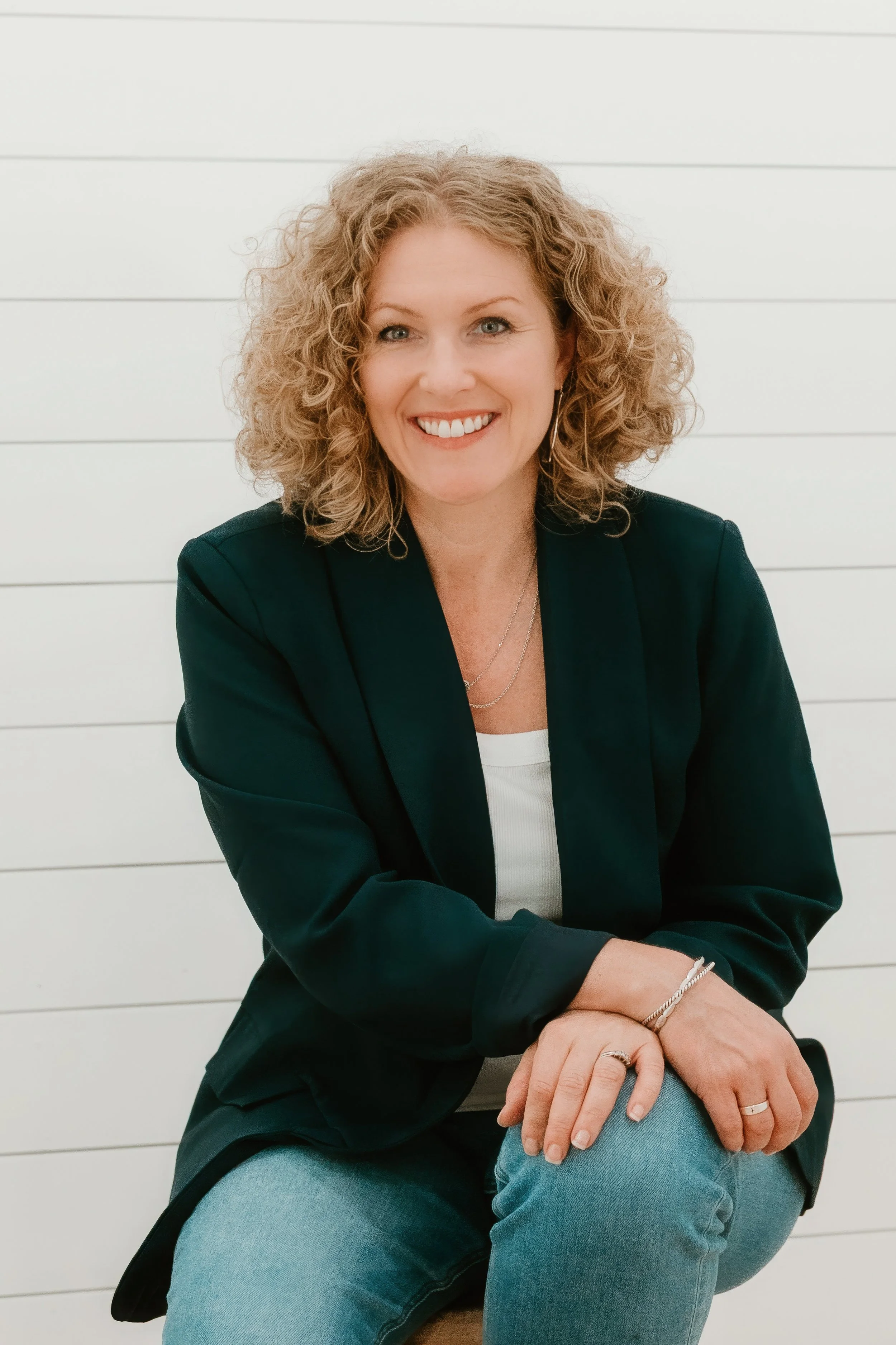 A woman with curly blond hair smiling, wearing a black blazer, white top, and blue jeans, sitting against a white paneled wall.