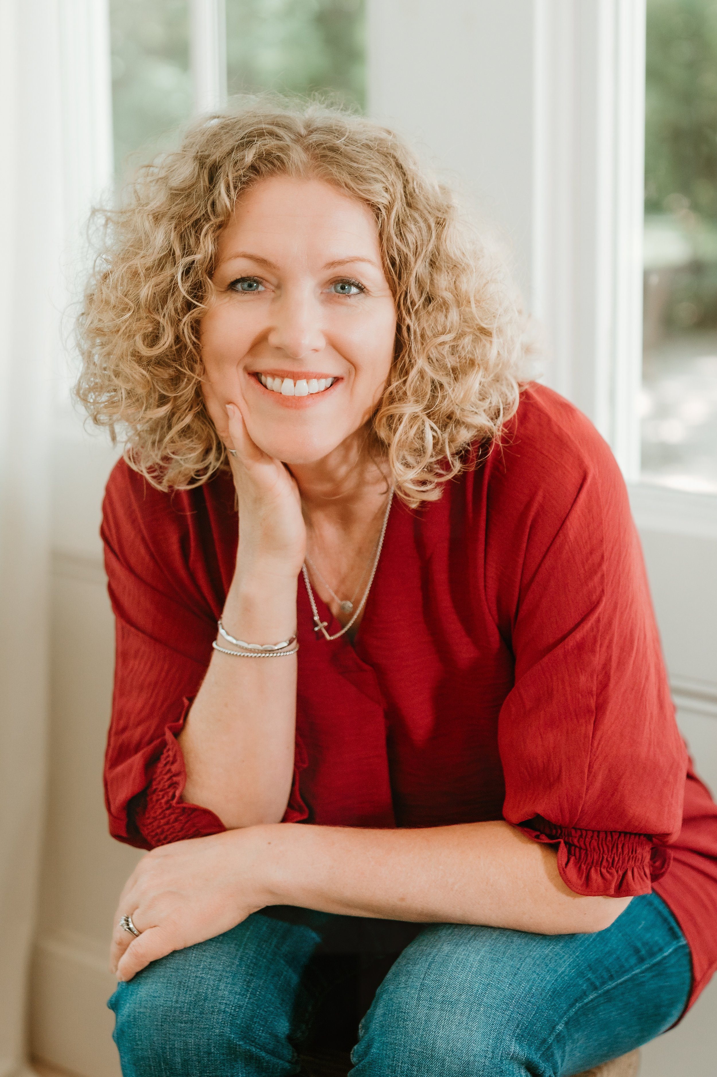 Portrait of a woman with curly blonde hair wearing a red top and jeans, sitting indoors near a window, smiling at the camera.